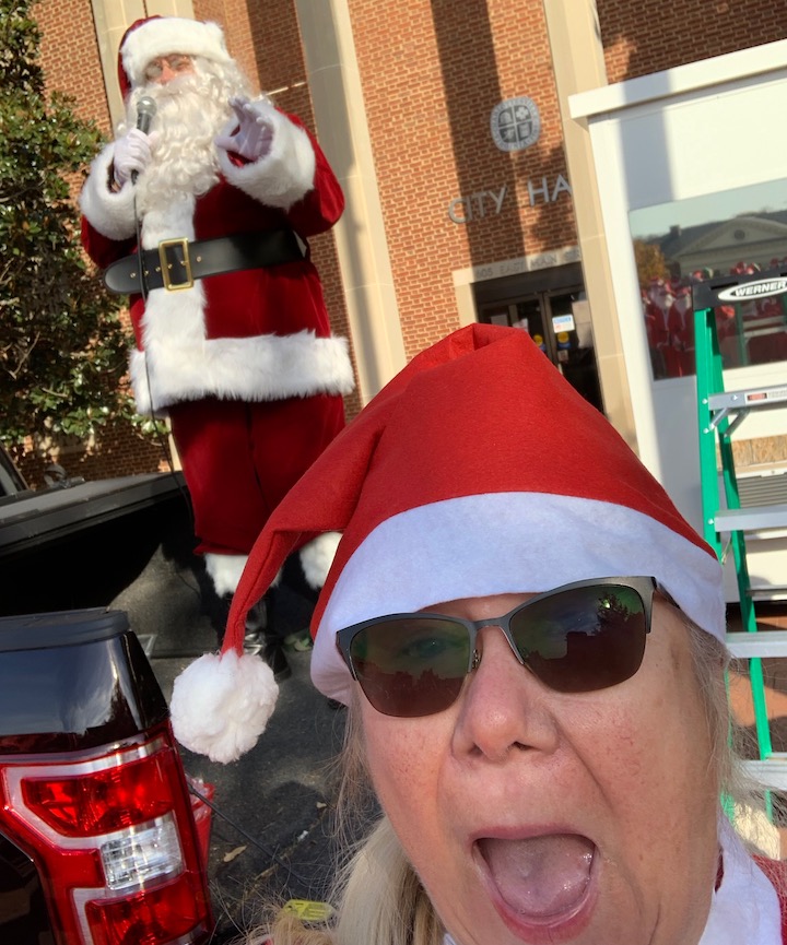 Person wearing a Santa hat and sunglasses, with Santa Claus in the background outside a building.