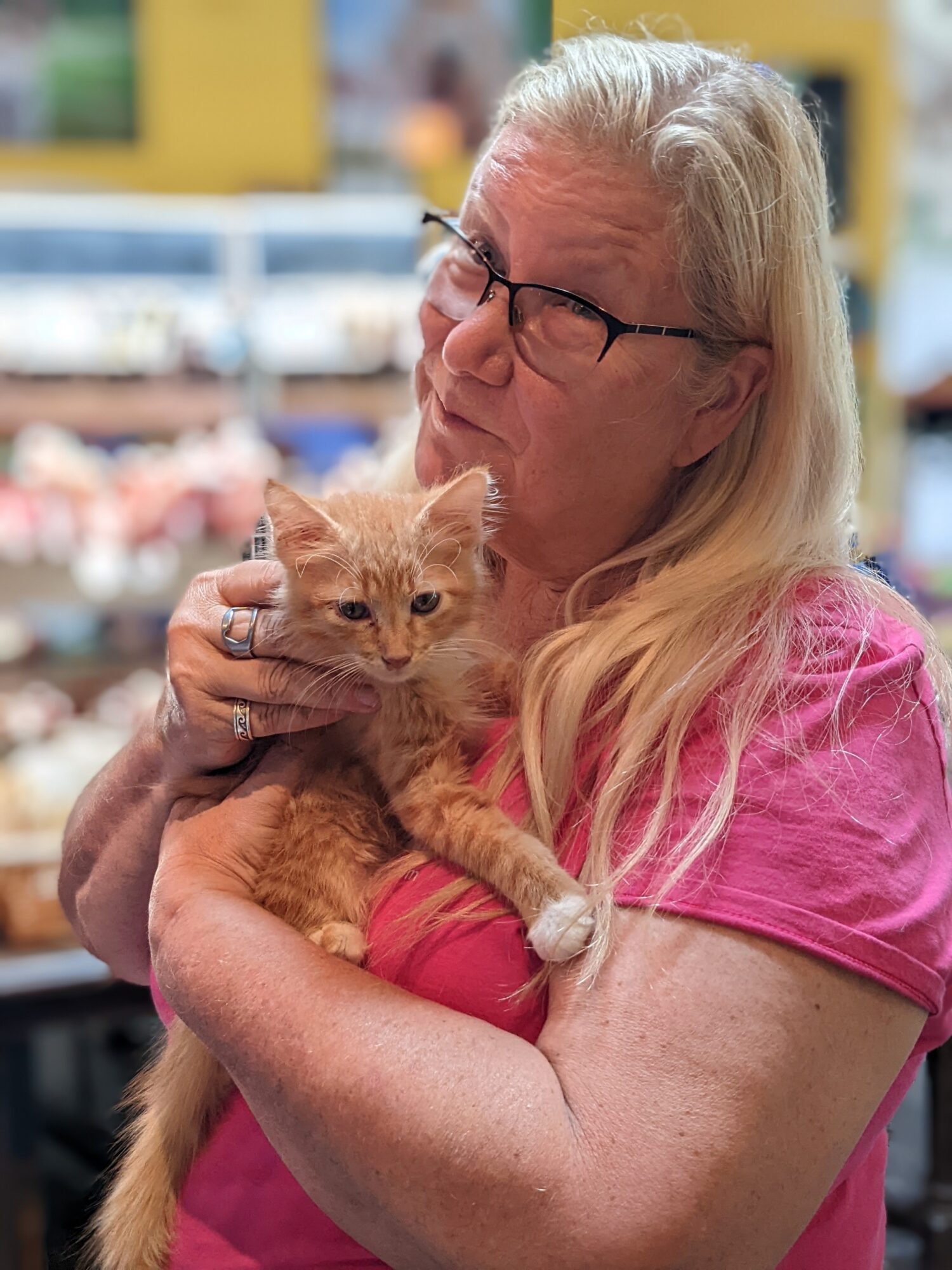 Older woman with glasses holding an orange kitten in a store, background blurred, woman wearing pink shirt, long blonde hair.
