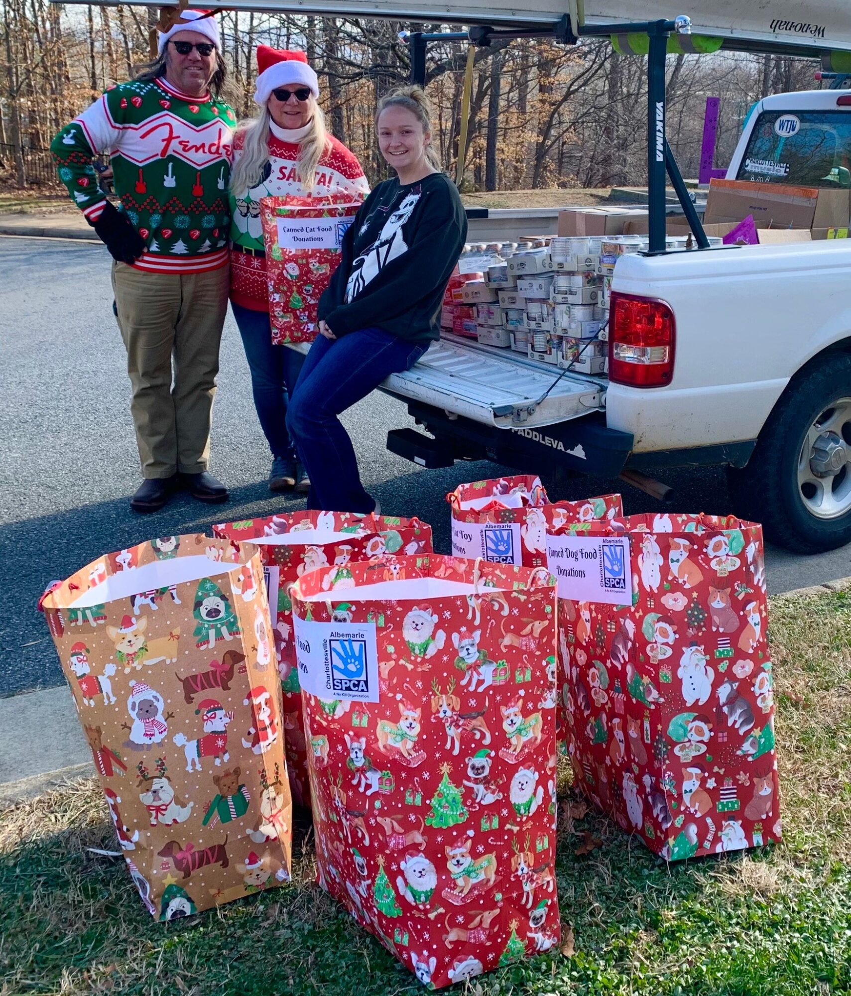 Three people stand next to a pickup truck with wrapped Christmas presents in the bed and on the ground, outdoors.