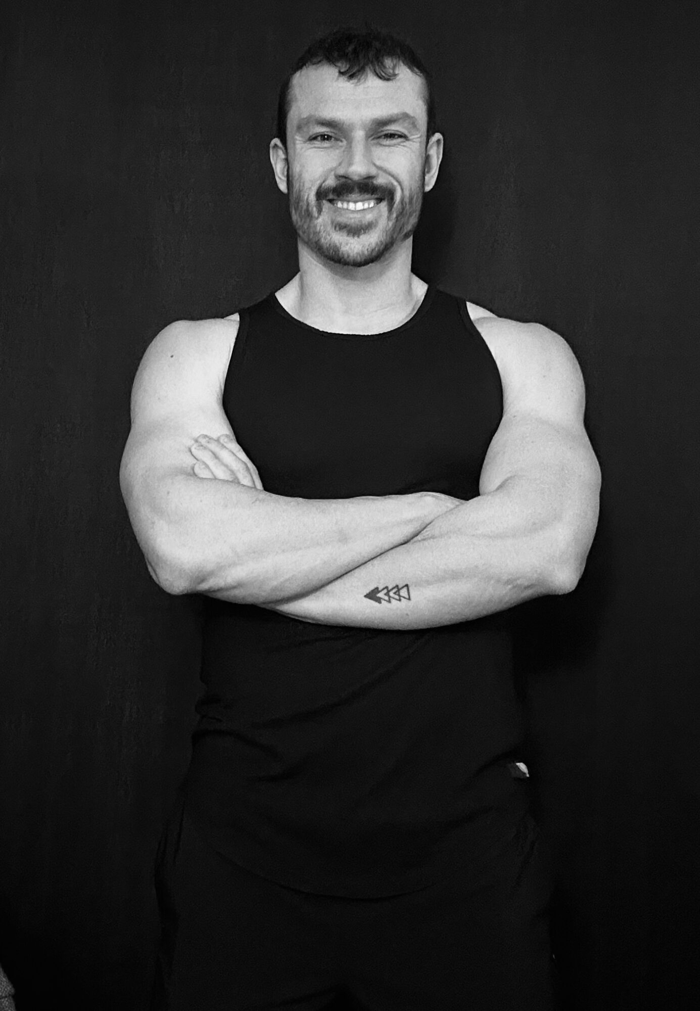 Man with short hair and beard smiling, arms crossed, wearing a sleeveless black shirt, standing against a dark background.