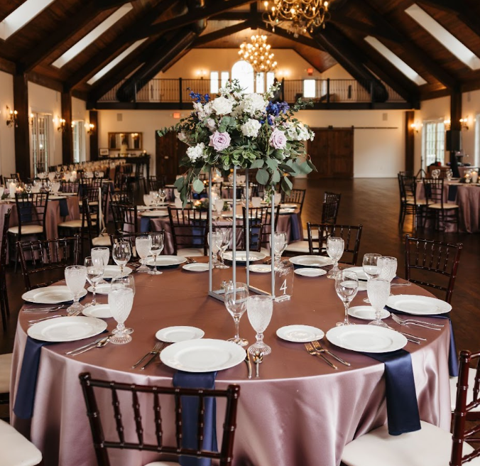 Round table with pink tablecloth, set with white plates, glasses, and silverware, decorated with a tall floral centerpiece, in a banquet hall.