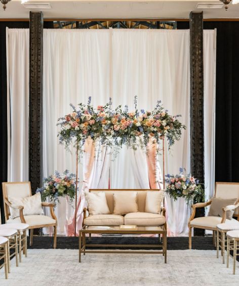Indoor wedding setup with a floral arrangement on a stand, cream-colored sofa, and chairs, with a white curtain backdrop.