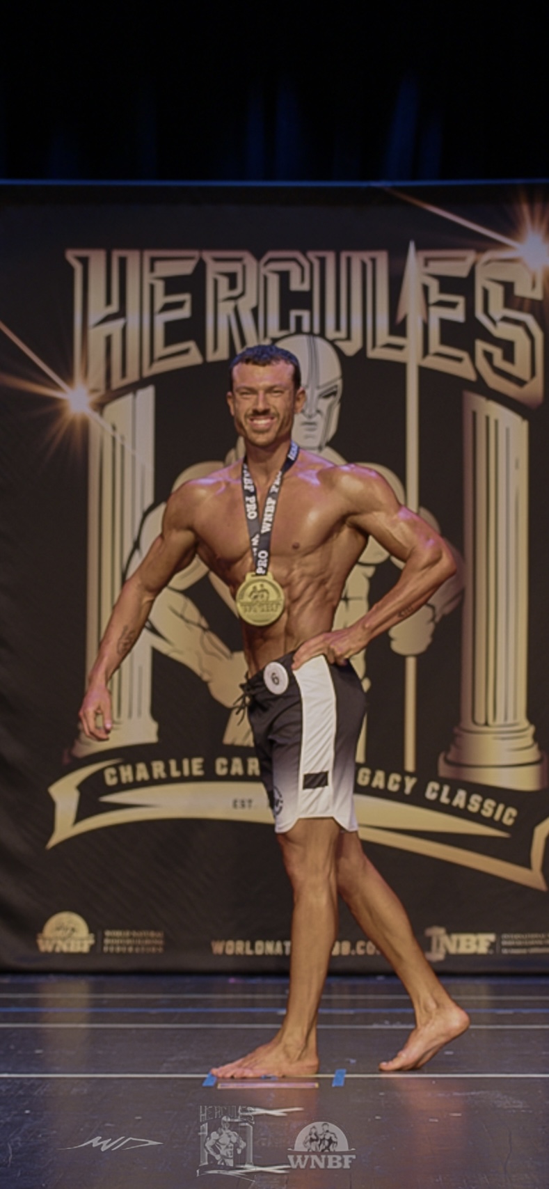 Man with a medal around his neck poses on stage in front of a Hercules-themed backdrop, wearing shorts and no shirt.