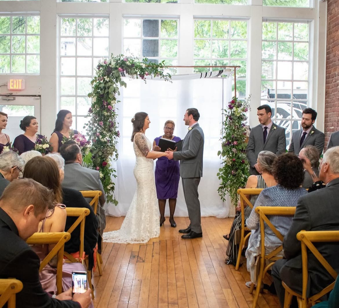 Bride and groom exchanging vows under floral arch, guests seated indoors with large windows, wooden floor, and natural light.