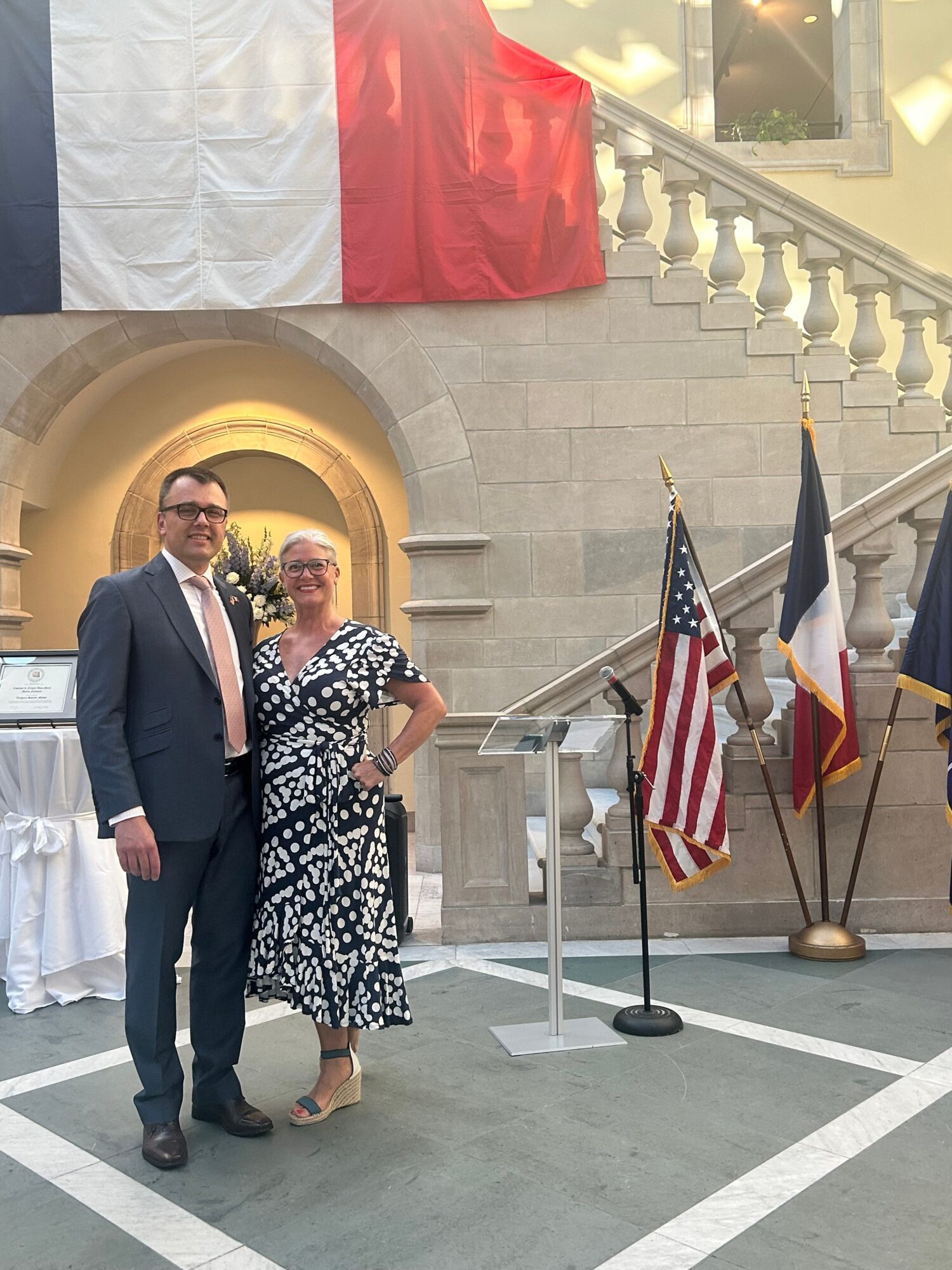 Two people standing in front of a flag display, one in a suit and the other in a patterned dress, with flags and stairs in background.