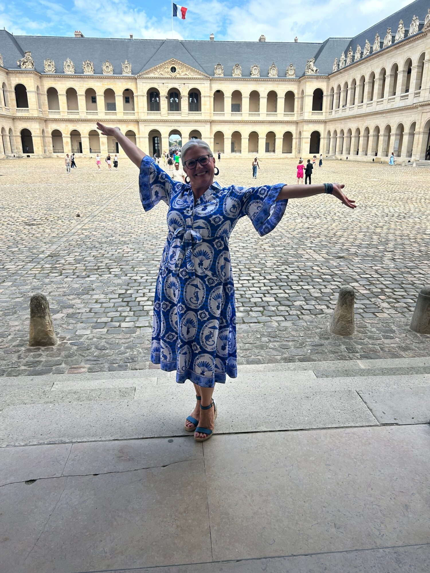 Person in a blue patterned dress standing with arms outstretched in front of a historic building, cobblestone ground, cloudy sky.