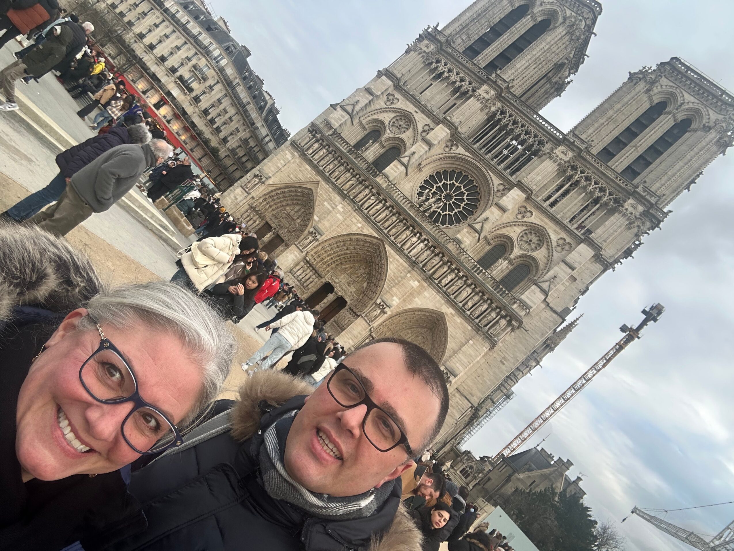 Two people with glasses smiling in front of Notre-Dame Cathedral in Paris, with tourists and buildings in the background.