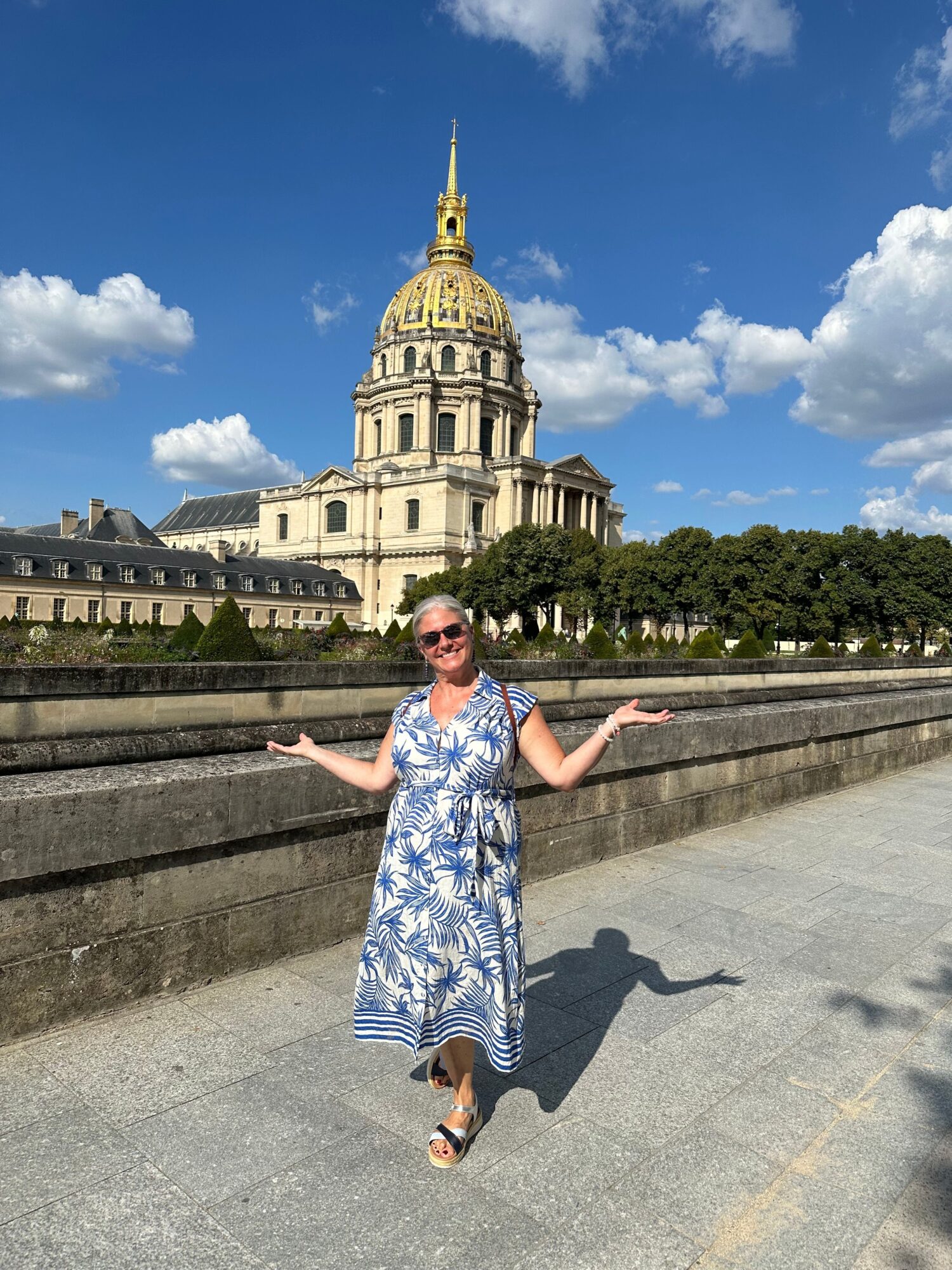 Woman in a blue and white dress poses on a bridge with a historic building and a golden-domed church in the background under a blue sky with clouds.