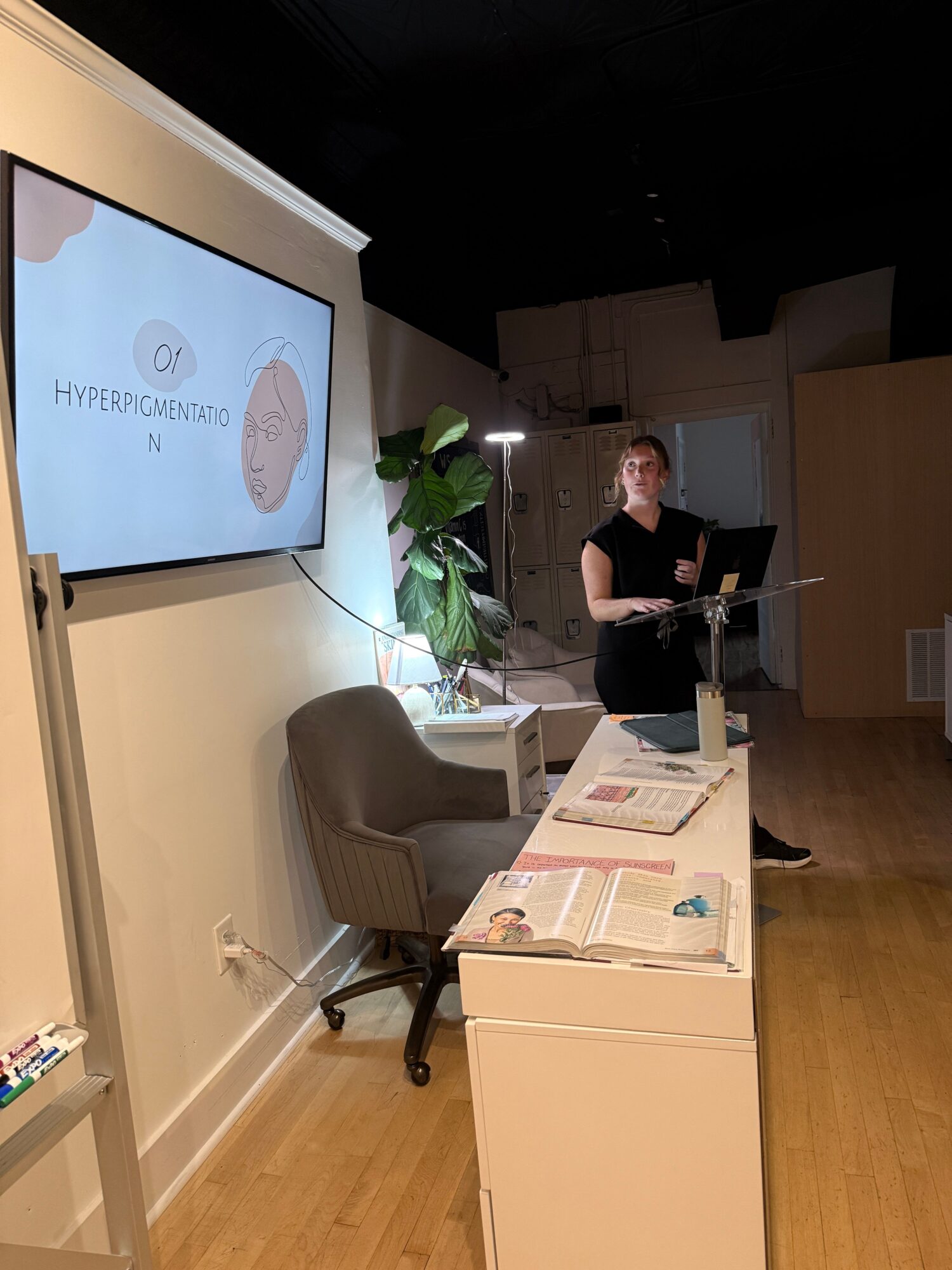 Person standing near a desk with papers, a plant, and a computer monitor in a room with wooden flooring.