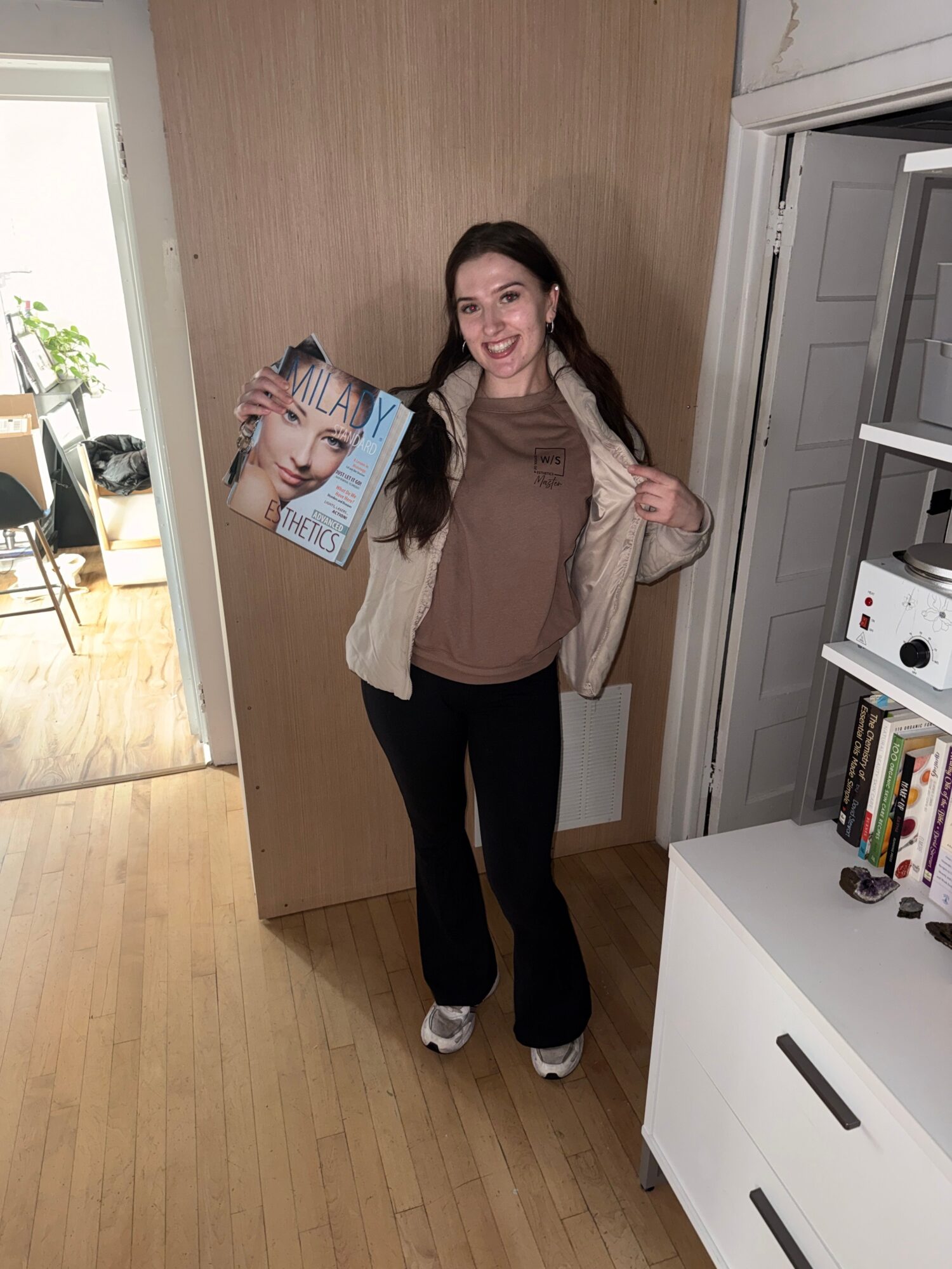 Woman standing in a room holding a magazine, smiling, with bookshelves and a window in the background.