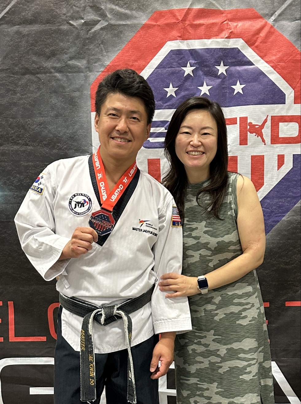 A man in a martial arts uniform with a medal and a woman in a sleeveless dress stand together, smiling in front of a banner.