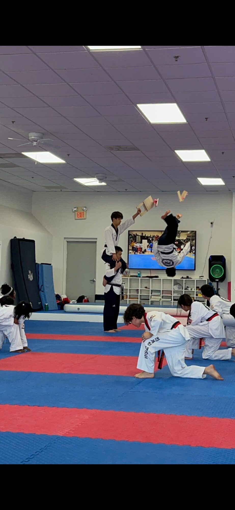 Martial arts class with students practicing on mats, instructor demonstrating techniques, and a screen in the background.