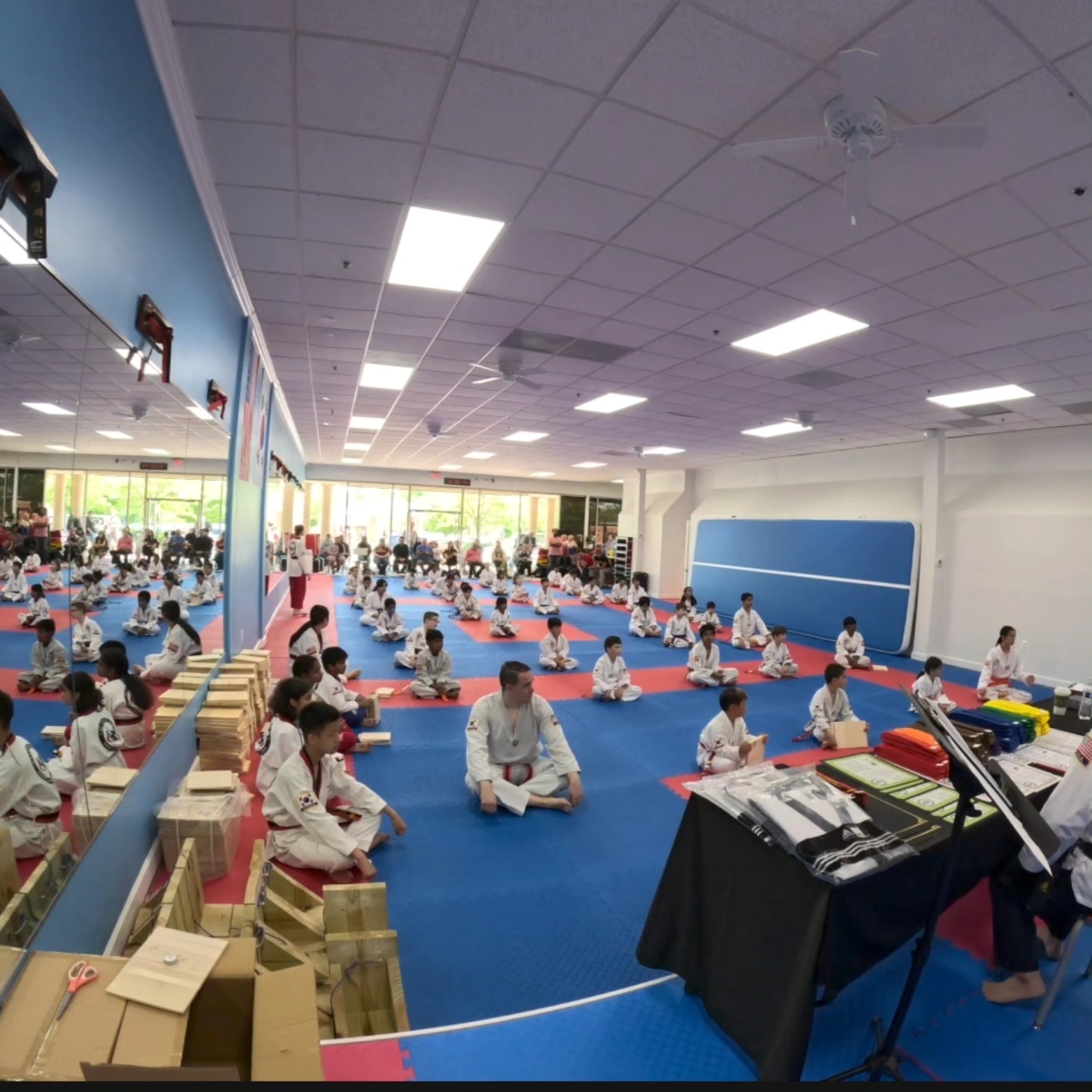 Martial arts class with children and instructor sitting on blue mats in a spacious room, practicing techniques, with equipment and mirrors visible.
