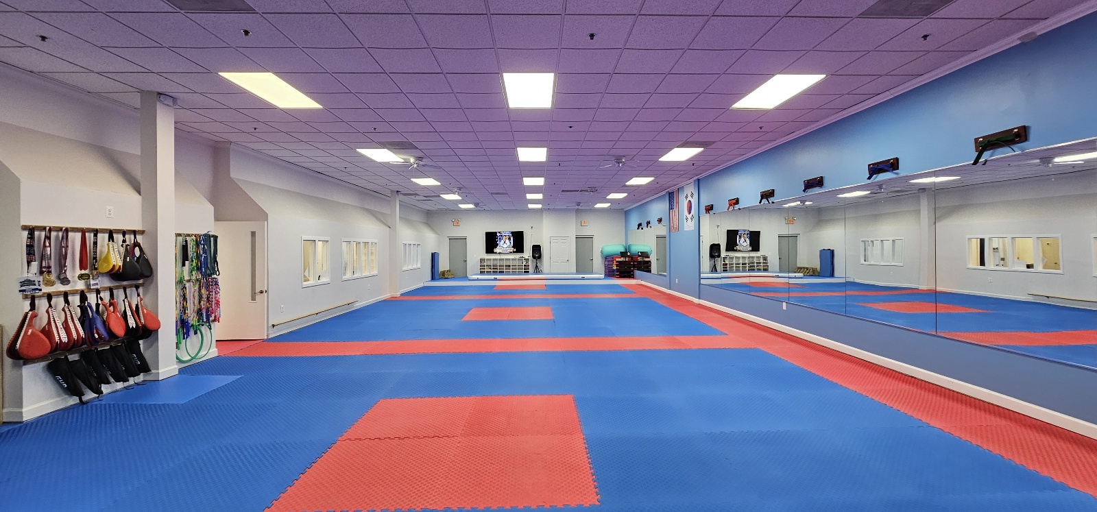 Empty martial arts training room with blue mats, red squares, mirrors, and equipment, well-lit with ceiling lights.