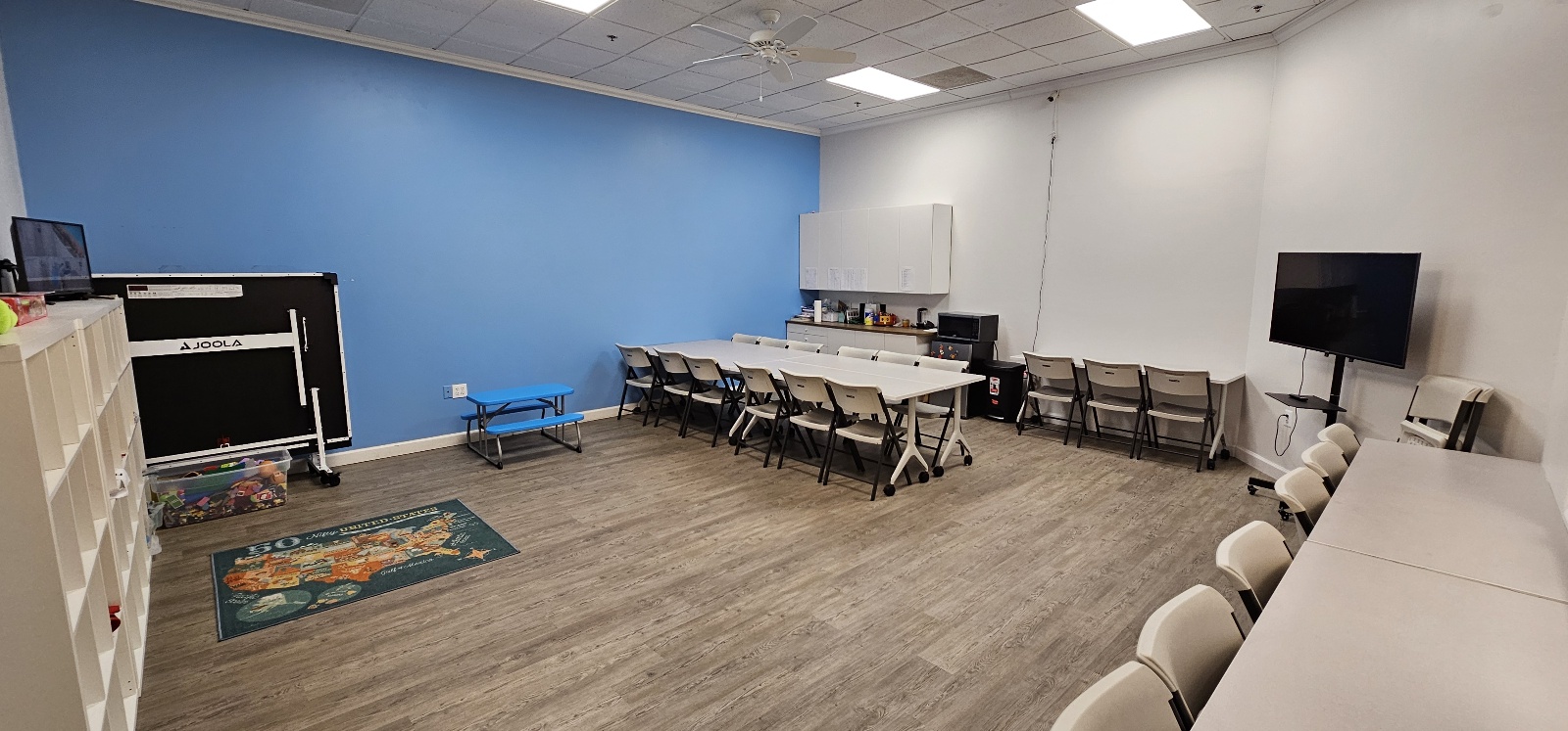 Empty room with a blue accent wall, tables, chairs, a TV, and a small rug, with a wooden floor.
