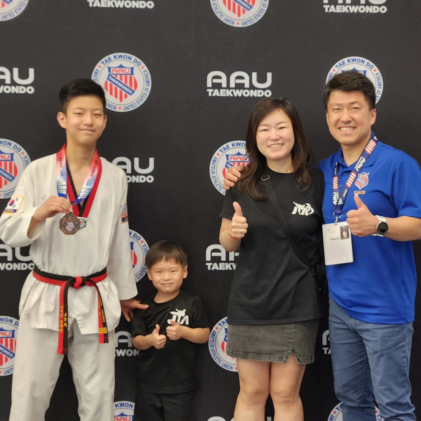 Group of four people, two children and two adults, smiling and giving thumbs up in front of a taekwondo backdrop.