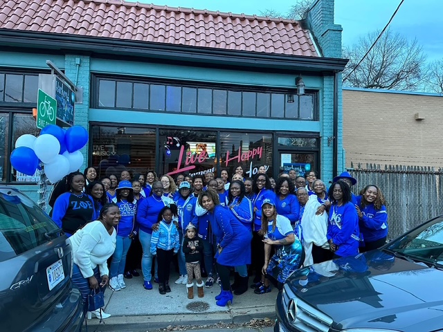 Group of people, including children, standing outside a store decorated with blue and white balloons, celebrating a birthday.