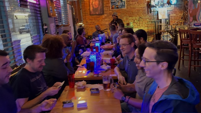 Group of people sitting at a long table in a lively bar or restaurant, engaging in conversation and using devices.
