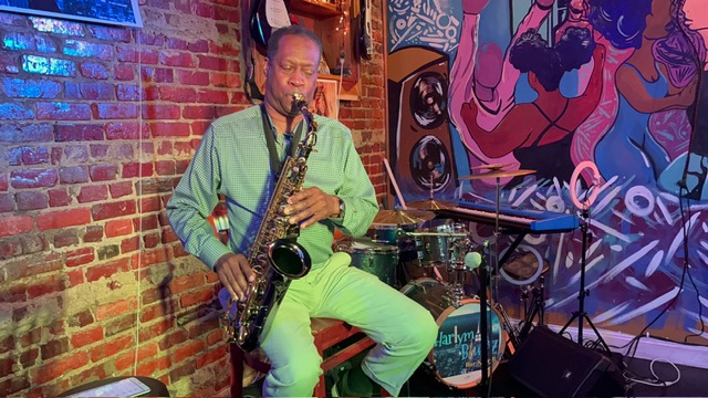 Person playing saxophone on stage with brick wall and colorful mural background, music equipment nearby.