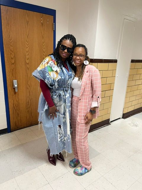 Two women standing indoors, one in pink plaid pajamas and the other in colorful clothing, smiling and posing together.