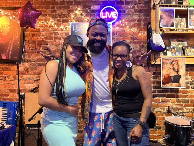 Three people smiling in a brick-walled room with musical instruments and neon signs, celebrating together.