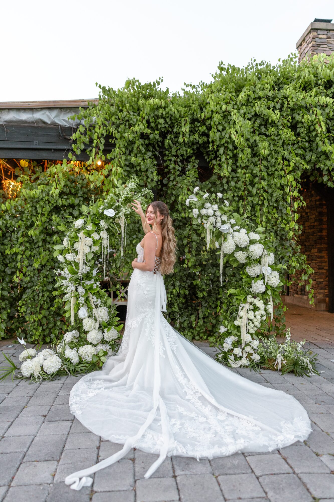 Woman in wedding dress standing in front of floral arch with greenery, on paved ground, outdoor setting