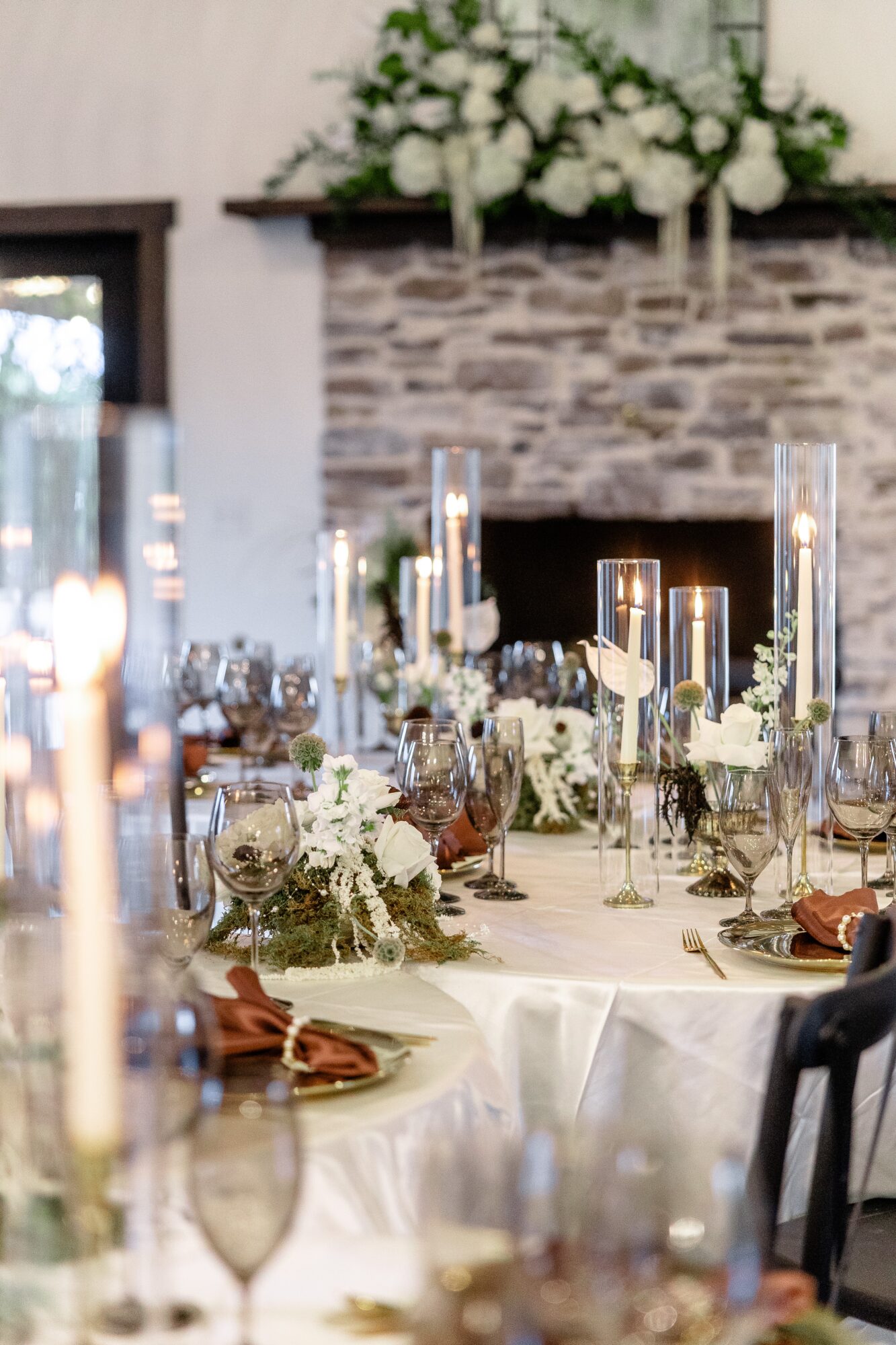 Elegant table setting with white flowers, candles, and glassware in a decorated room with a stone fireplace.