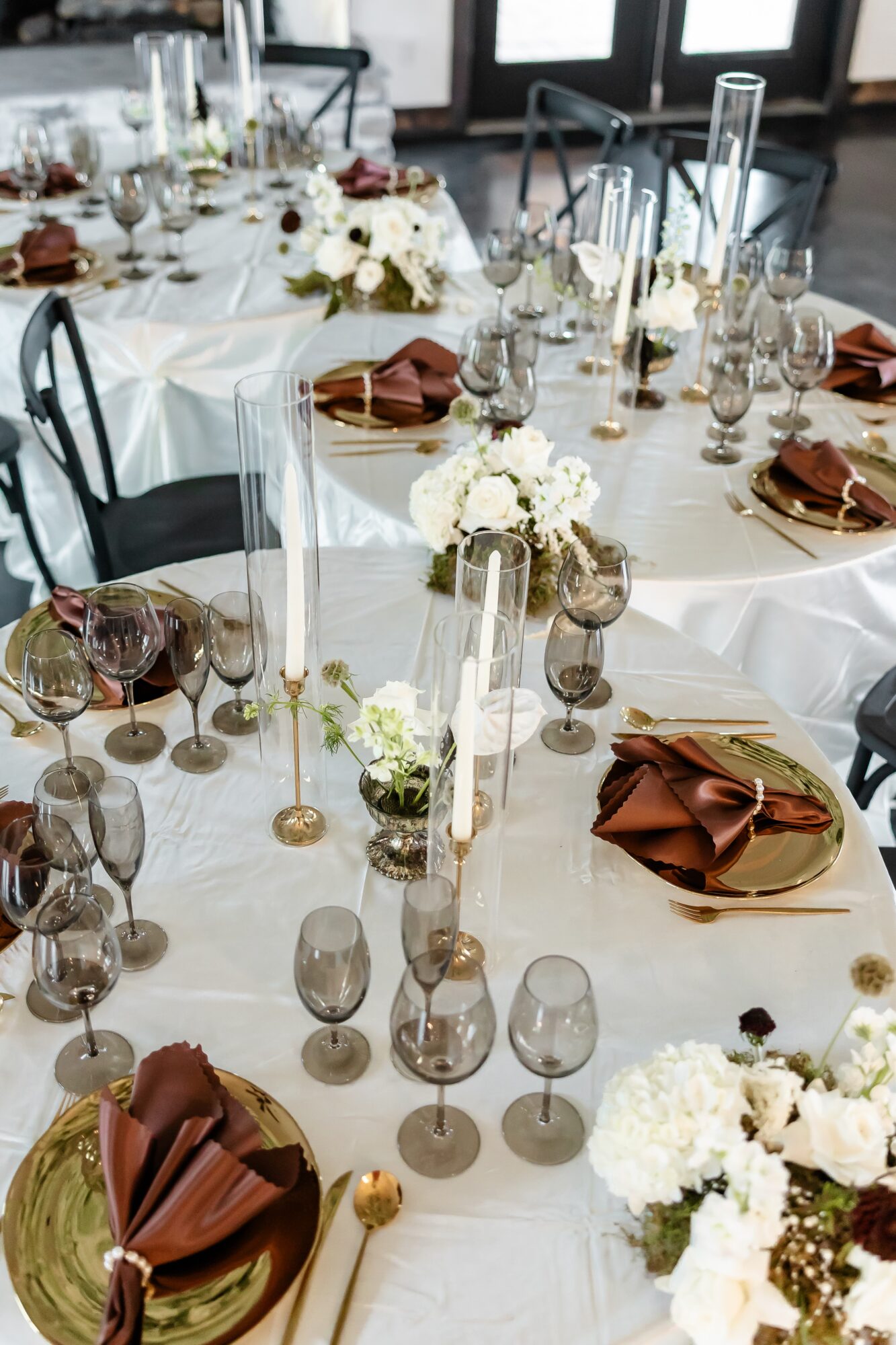 Elegant table setting with white flowers, candles, glassware, and brown napkins on a round table.