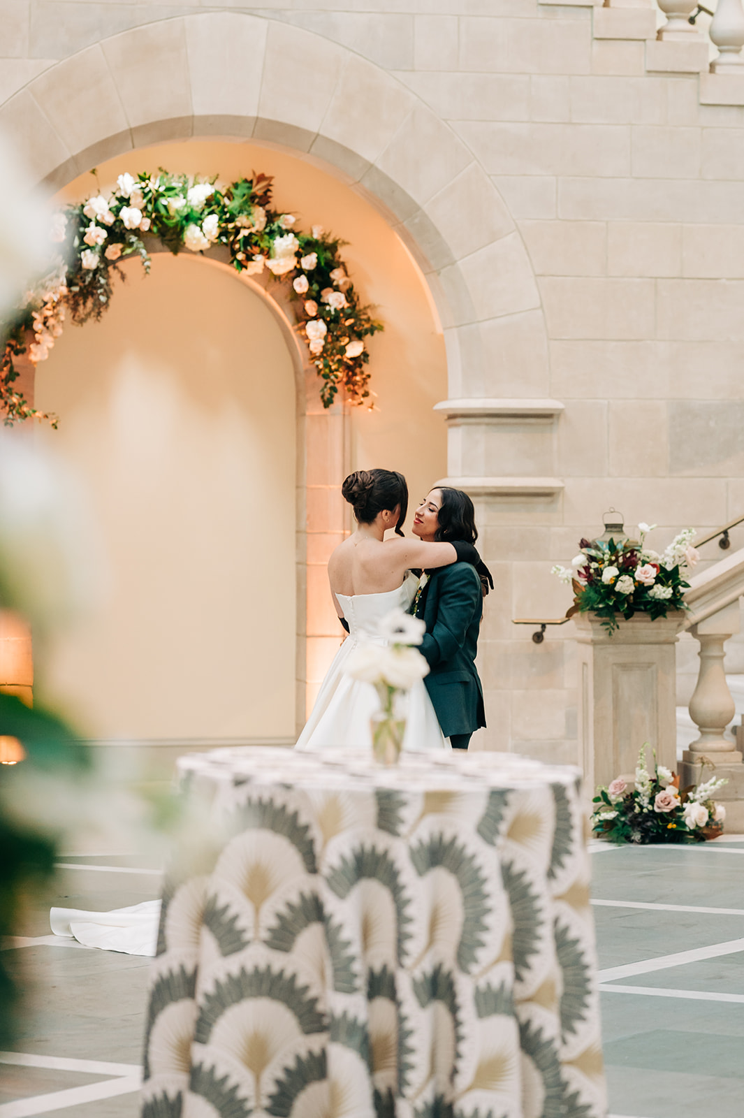 Two women in wedding attire embrace in front of an arch decorated with flowers, inside a stone building.