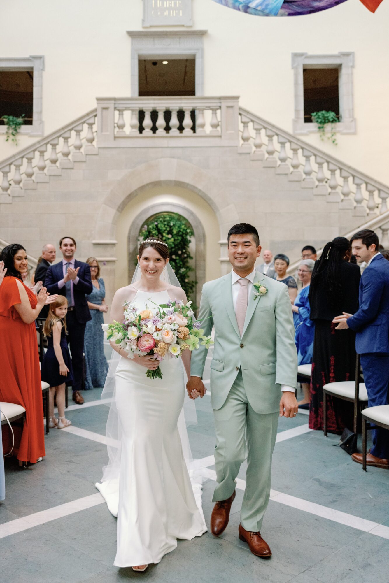 Bride and groom walking hand in hand outside a building with stairs, surrounded by guests, bride holding a bouquet, groom in light suit.