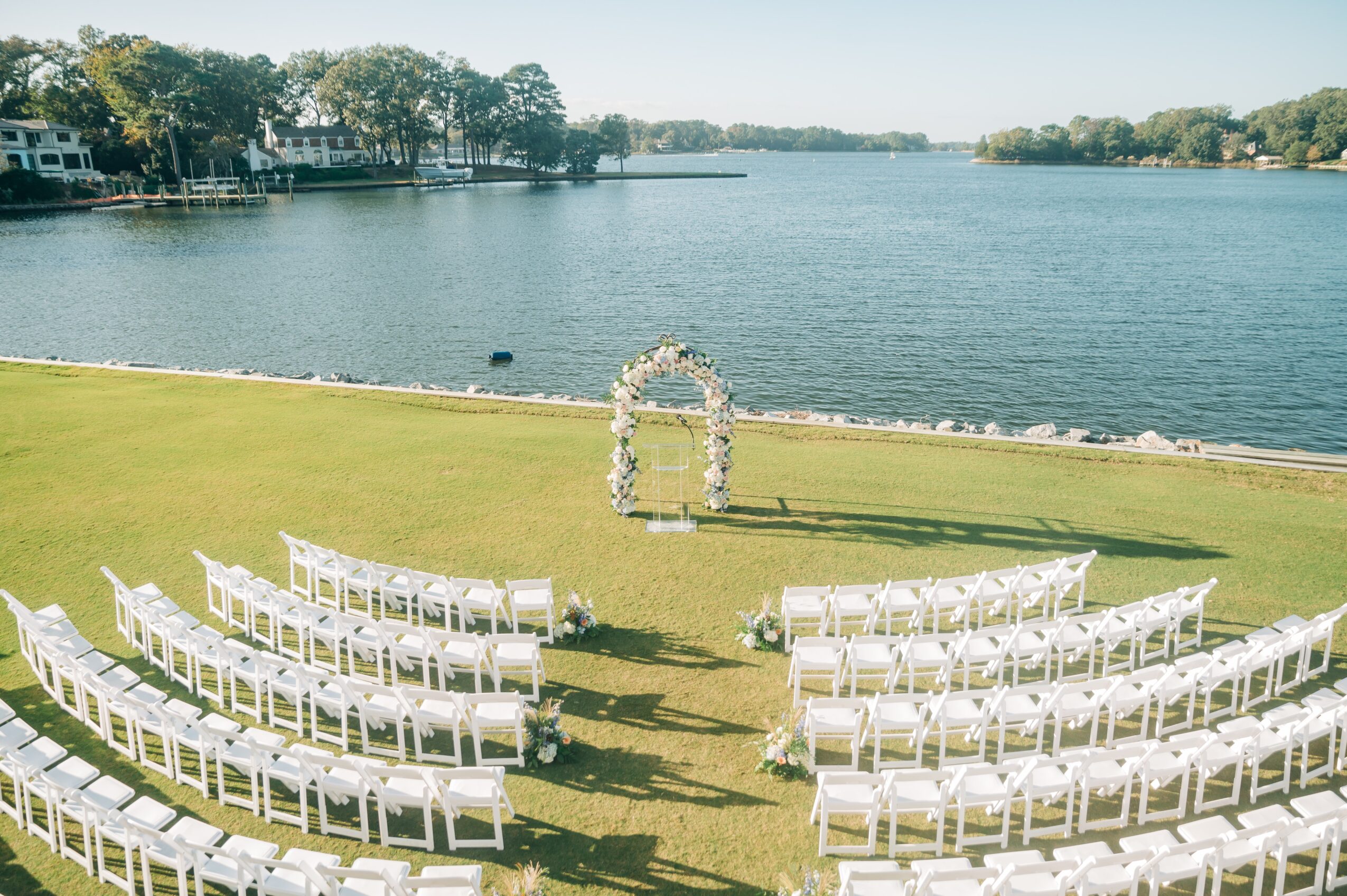 Outdoor wedding setup with white chairs facing a lake, decorated arch, and clear sky.