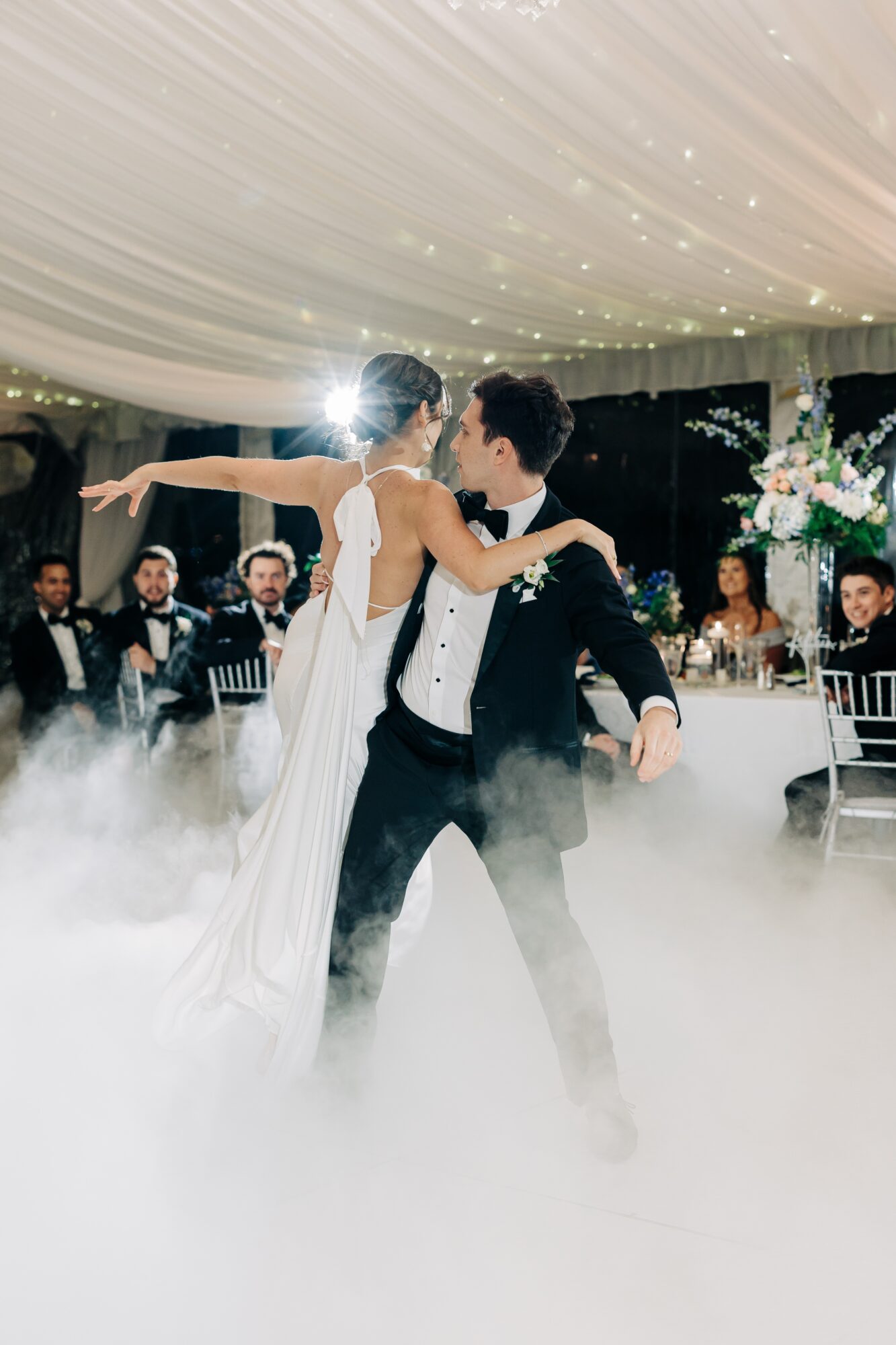 A bride and groom dance under a decorated ceiling with guests seated at tables, surrounded by fog and soft lighting.
