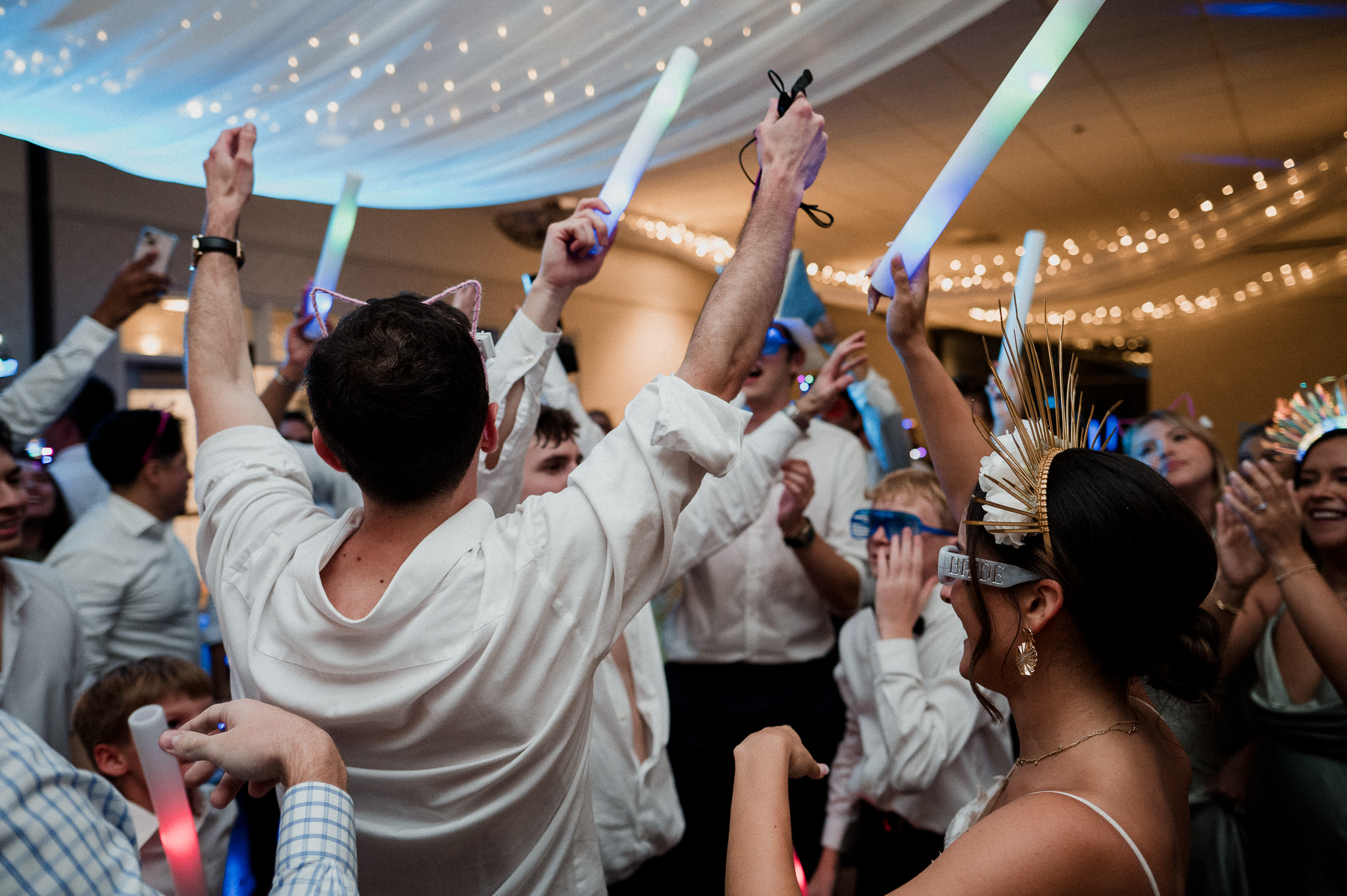People celebrating at a party, holding glow sticks and raising hands, with a decorated ceiling and string lights in the background.