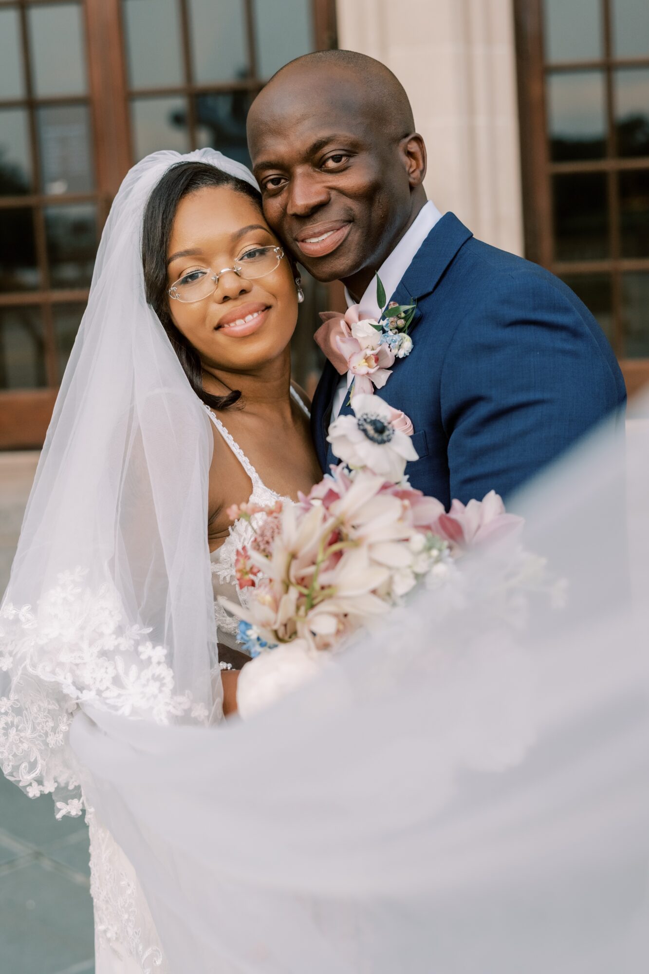 A bride and groom pose outdoors, smiling, with the bride holding a bouquet of flowers, in front of a building with large windows.