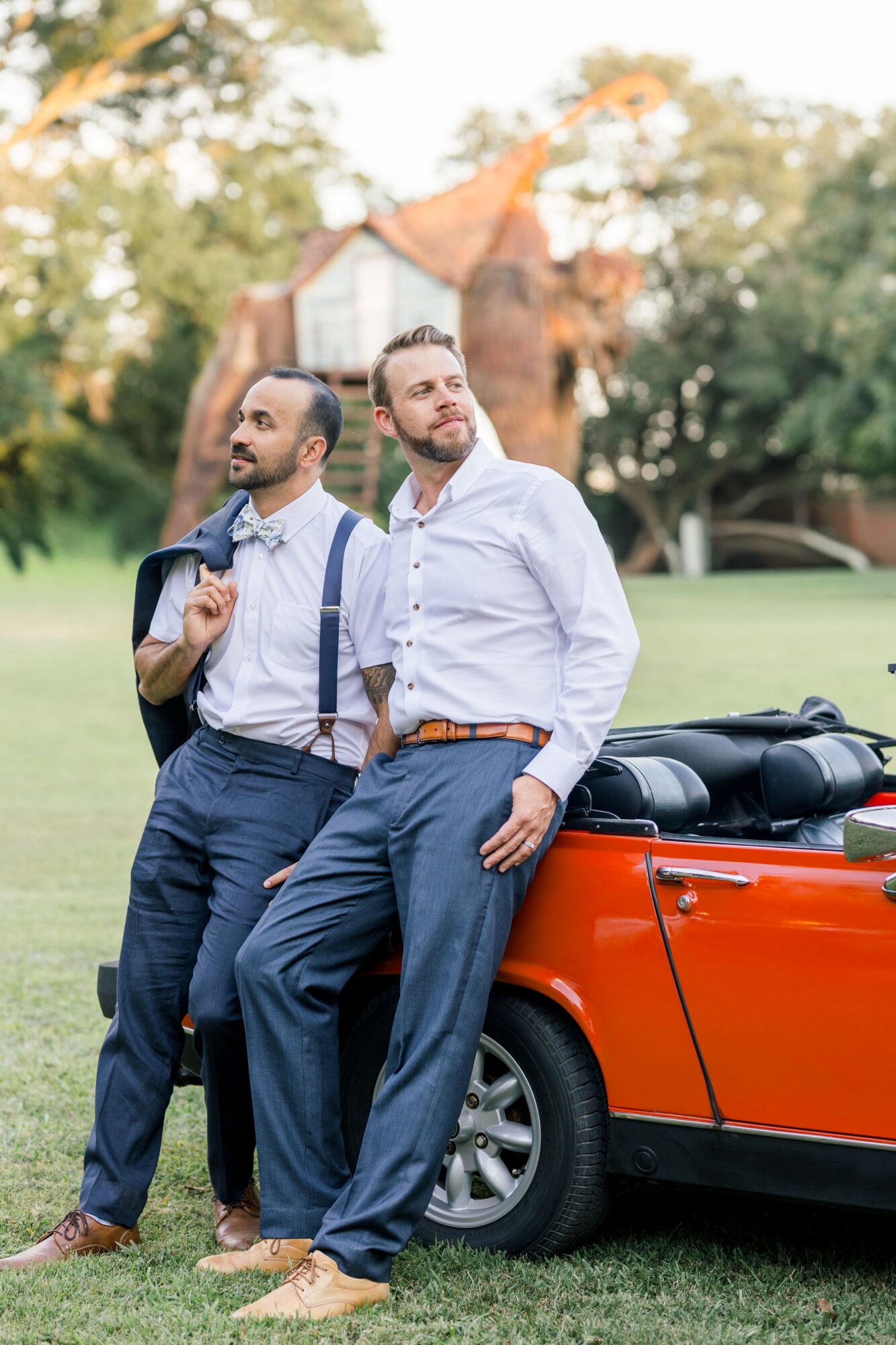 Two men in formal attire leaning against a red convertible car outdoors, with trees and a building in the background.