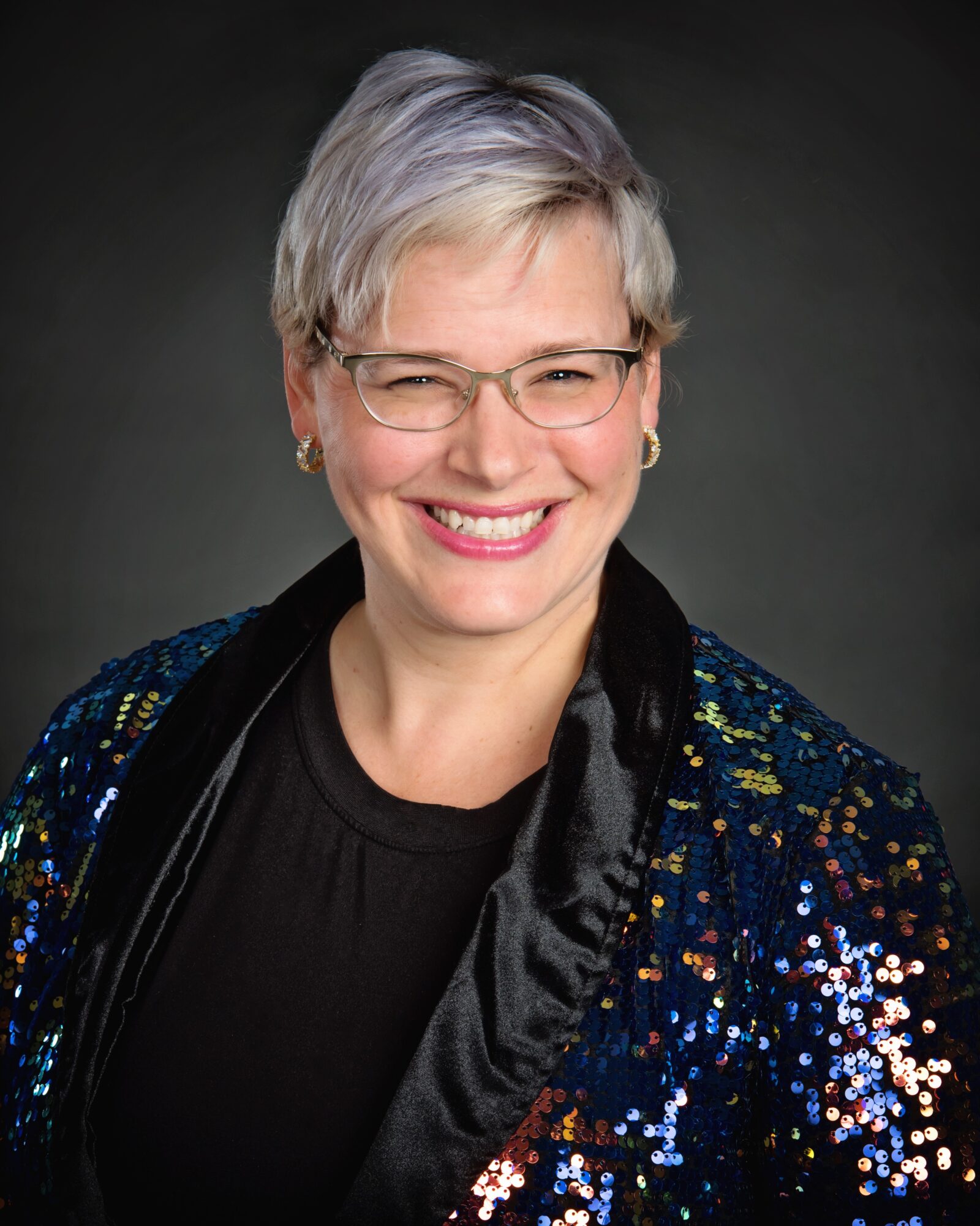 Smiling woman with short gray hair, glasses, wearing a black top and a sequined jacket.