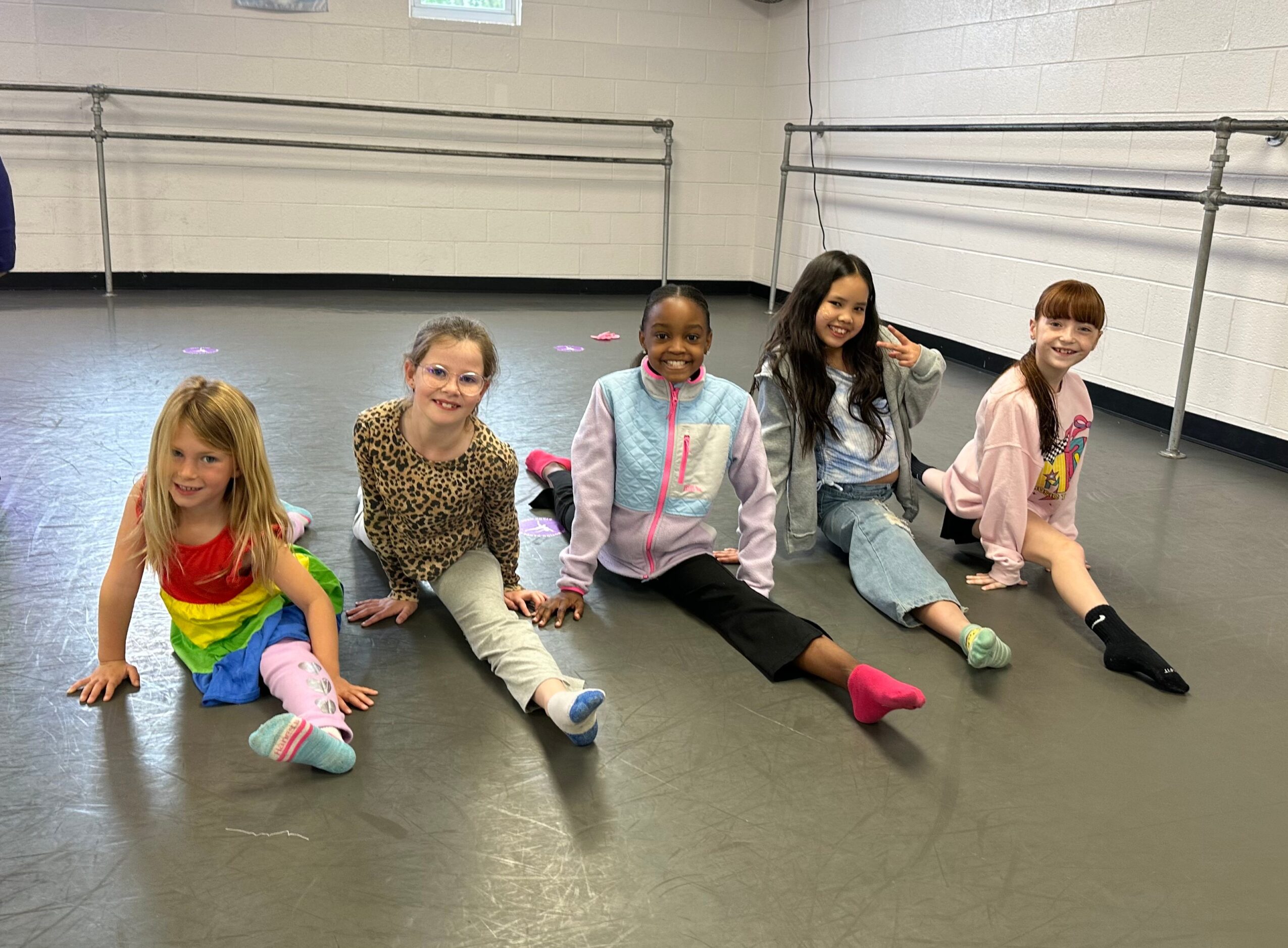 Five children sitting on the floor in a dance studio, smiling and posing for the camera.