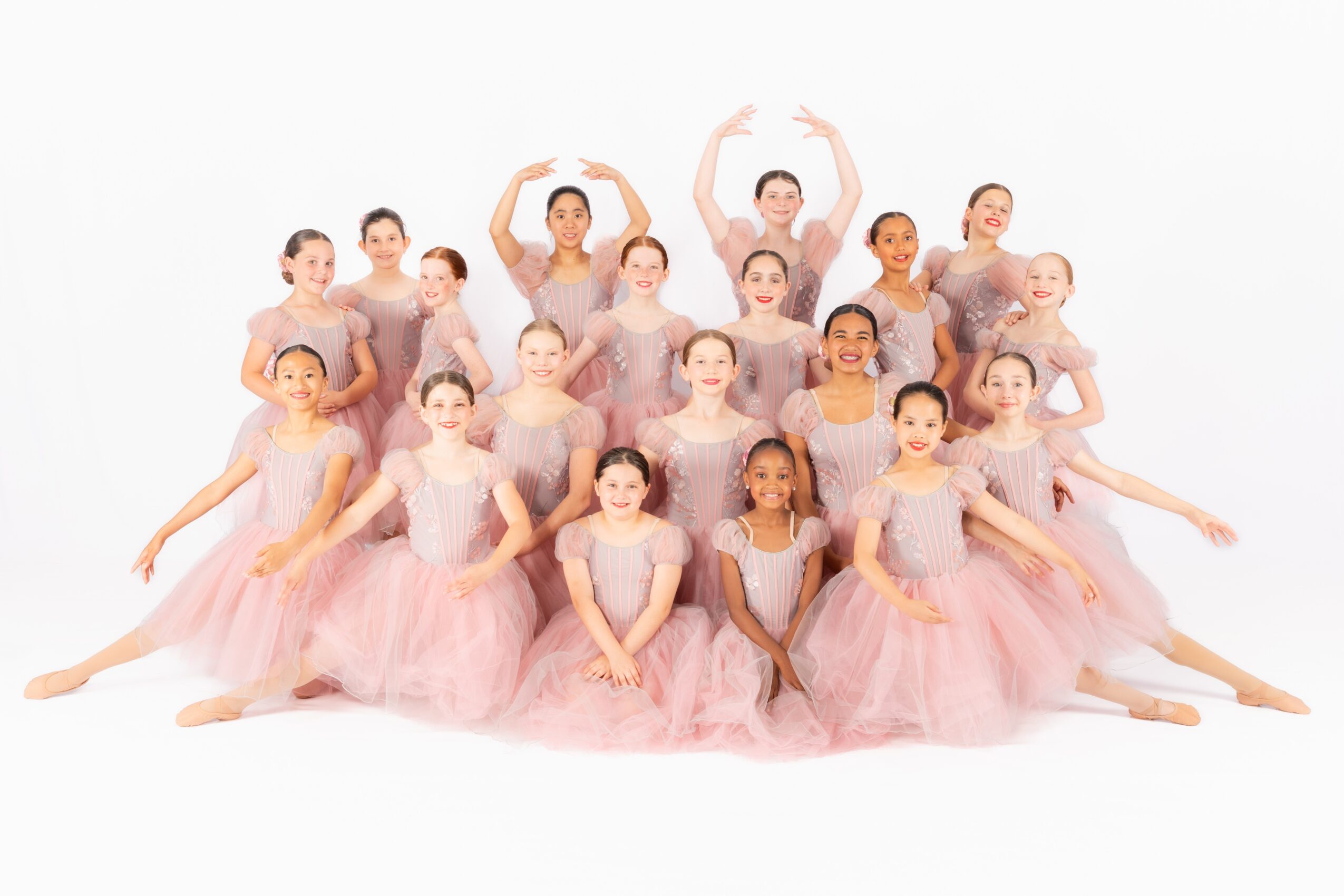 Group of young ballet dancers in pink tutus posing together, some sitting and some standing, on a white background.