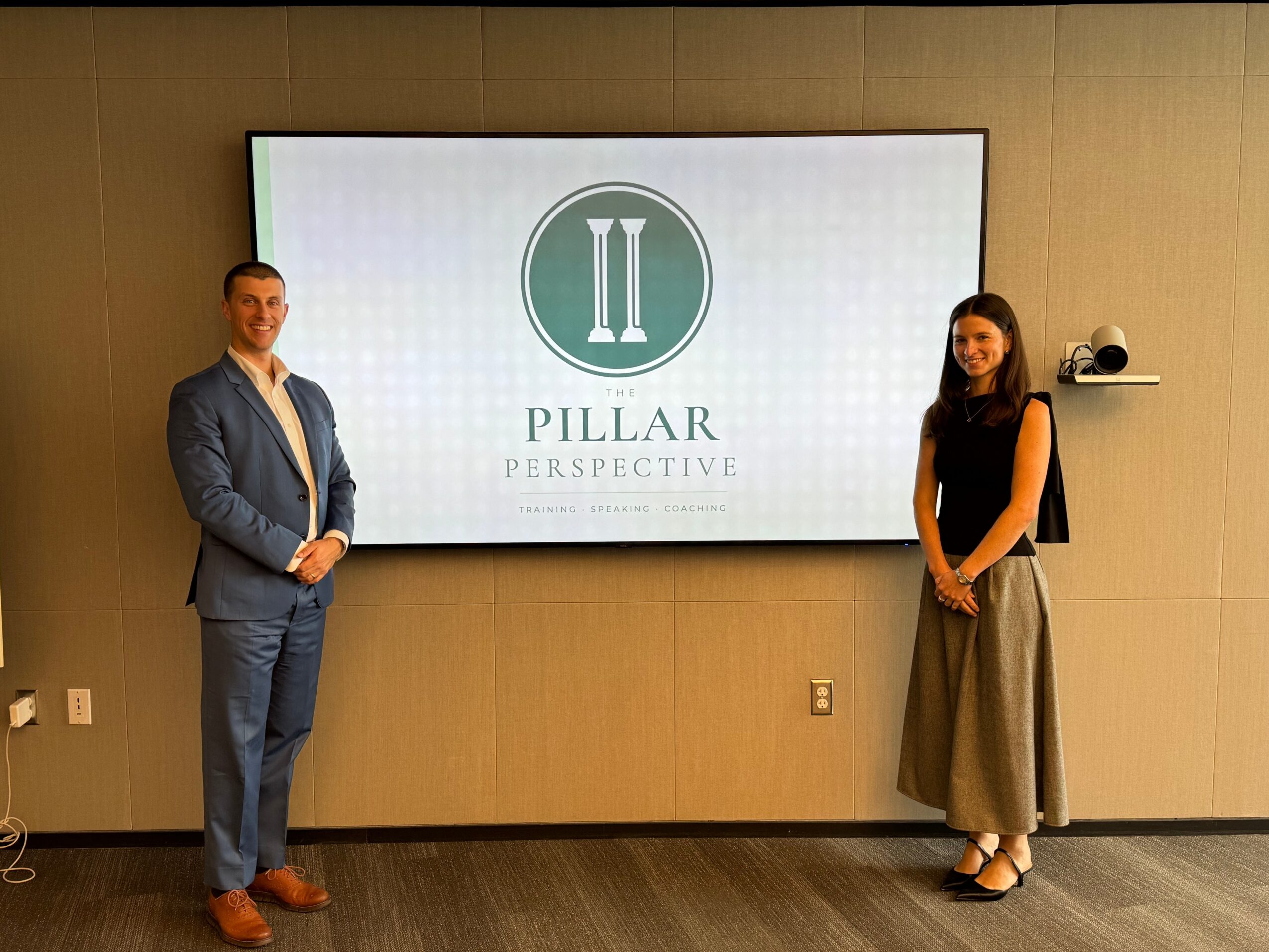 Two people standing in front of a large screen displaying a logo with pillars and the words 'Pillar Perspective'.