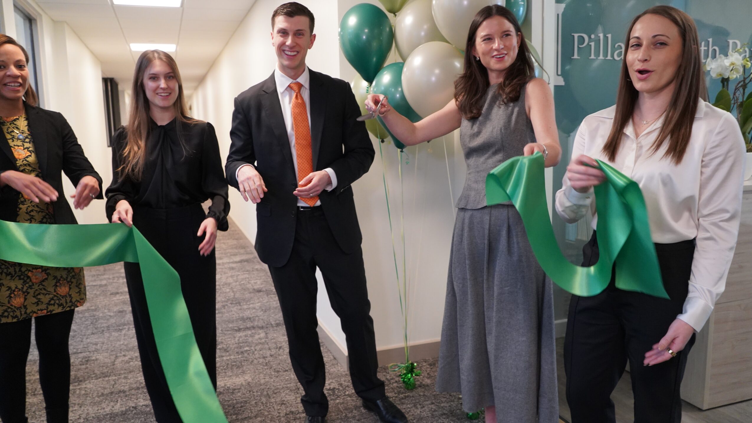 Group of five people at a ribbon-cutting event, holding a green ribbon, with balloons in the background.