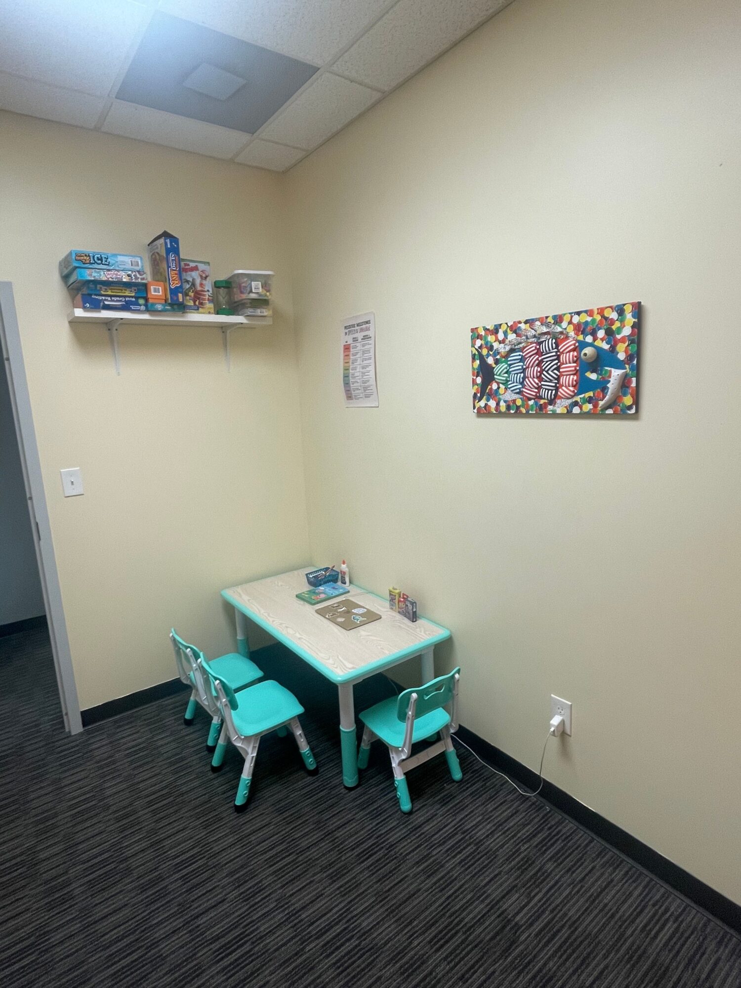 Small table with four chairs in a room corner, wall with colorful artwork, shelf with toys, and a window.