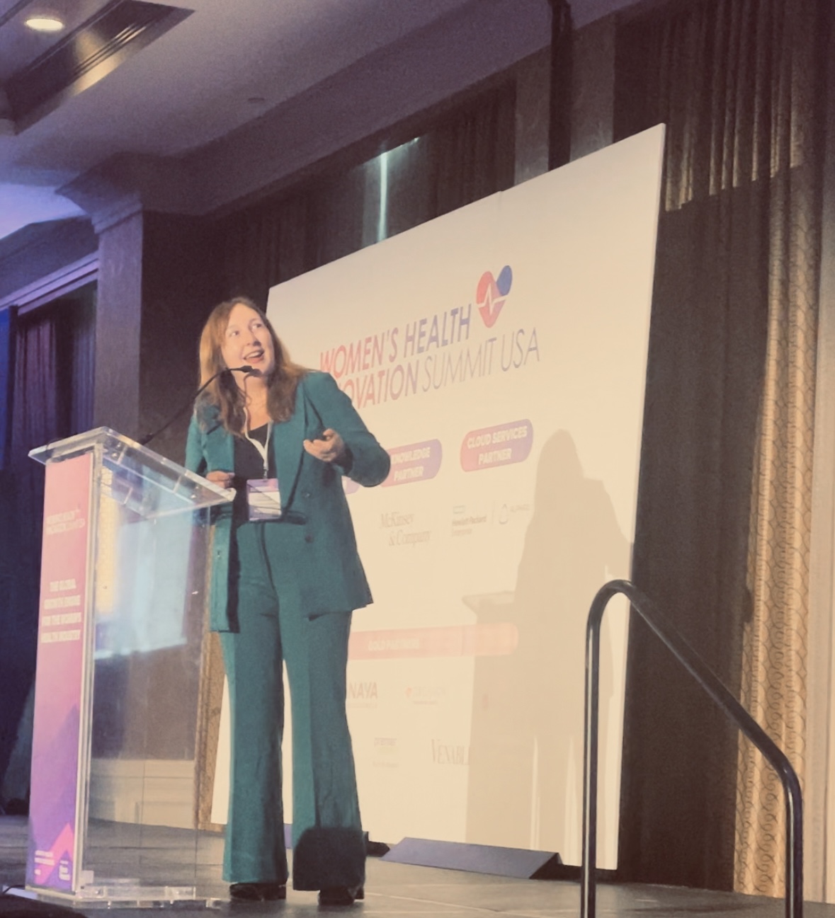 Woman speaking at a podium during a conference, with a banner behind her reading 'Women's Health' and 'U.S.A.'