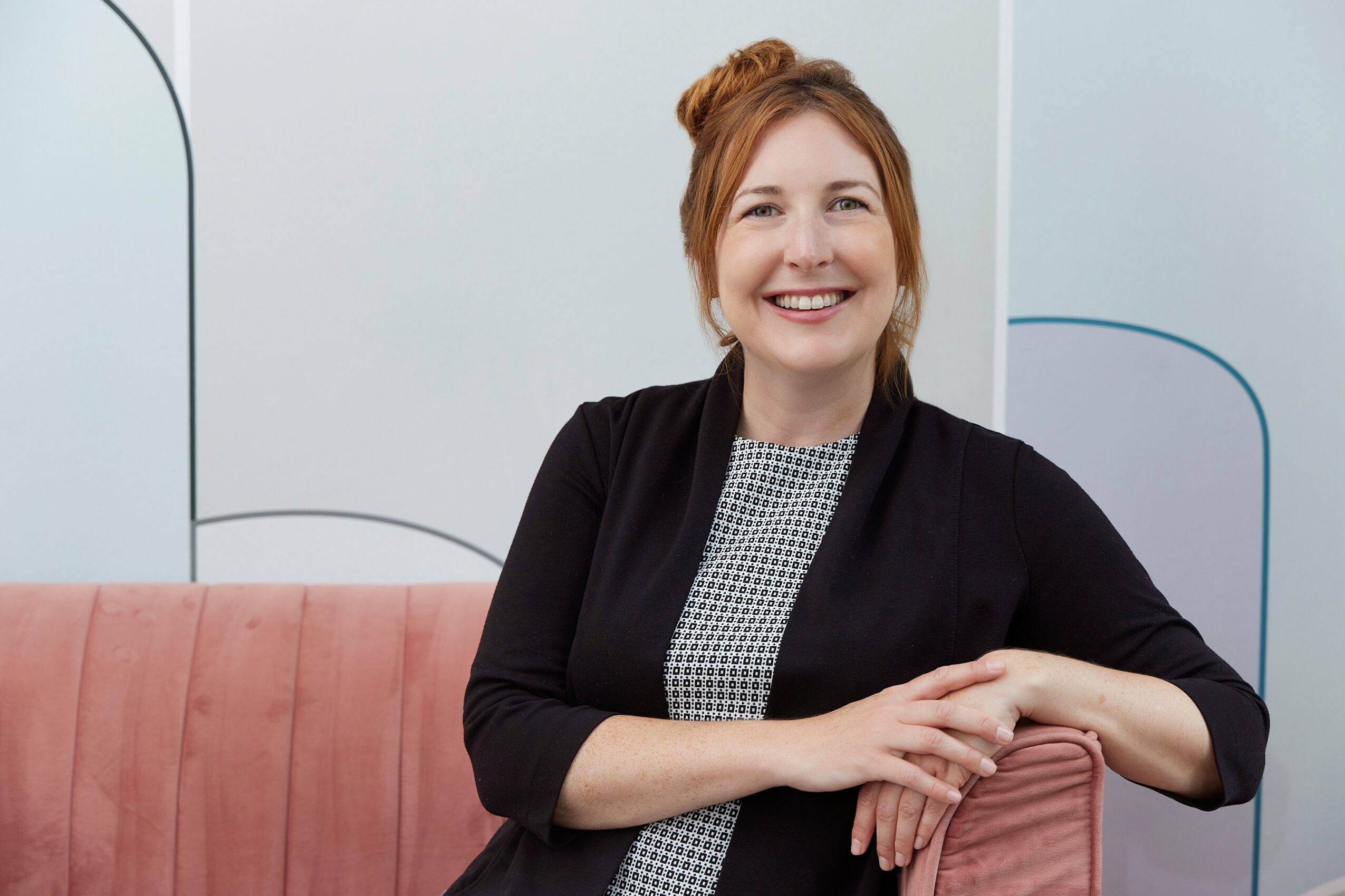 Smiling woman with red hair sitting on a pink couch in a modern room, wearing a black cardigan and patterned top.