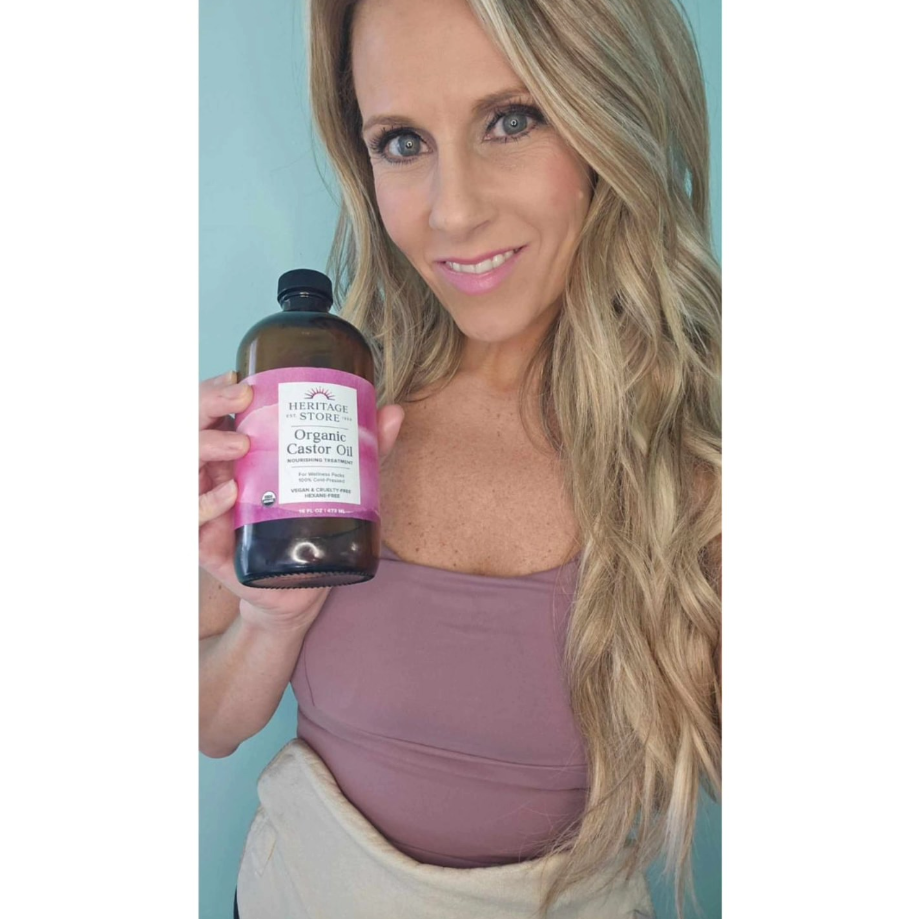 Woman with long blonde hair holding a bottle of organic coconut oil, smiling, wearing a mauve top and beige pants.