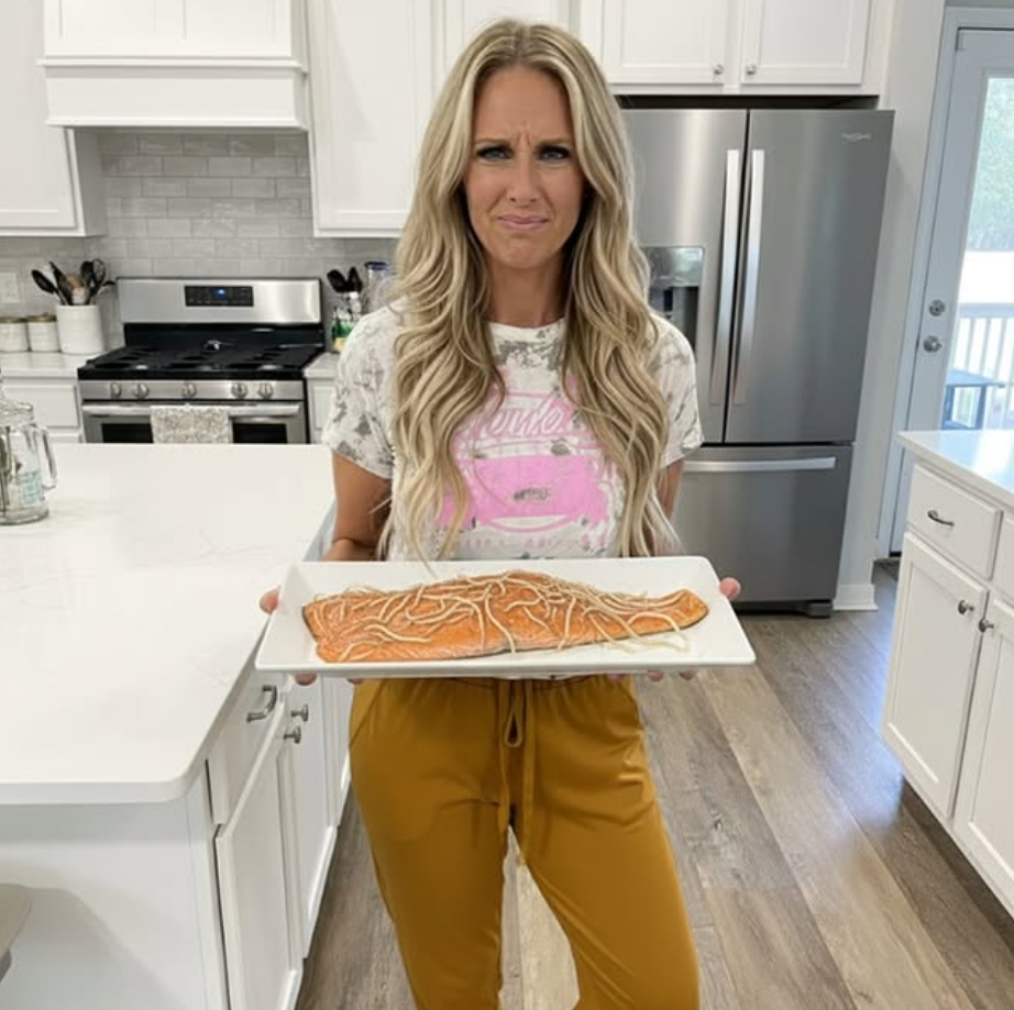 Woman holding a large fish on a white tray in a modern kitchen, with a confused expression, blonde hair, white cabinets, and stainless steel appliances.