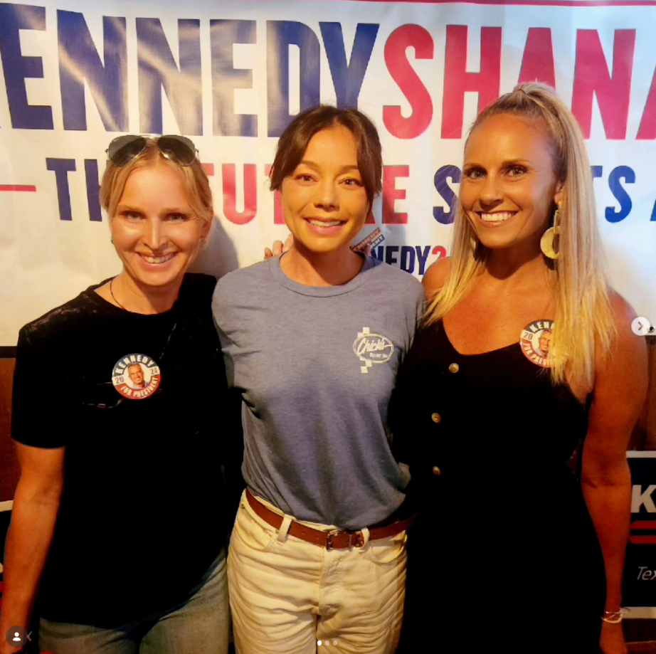 Three women standing together in front of a campaign sign, smiling, with campaign buttons on their shirts.