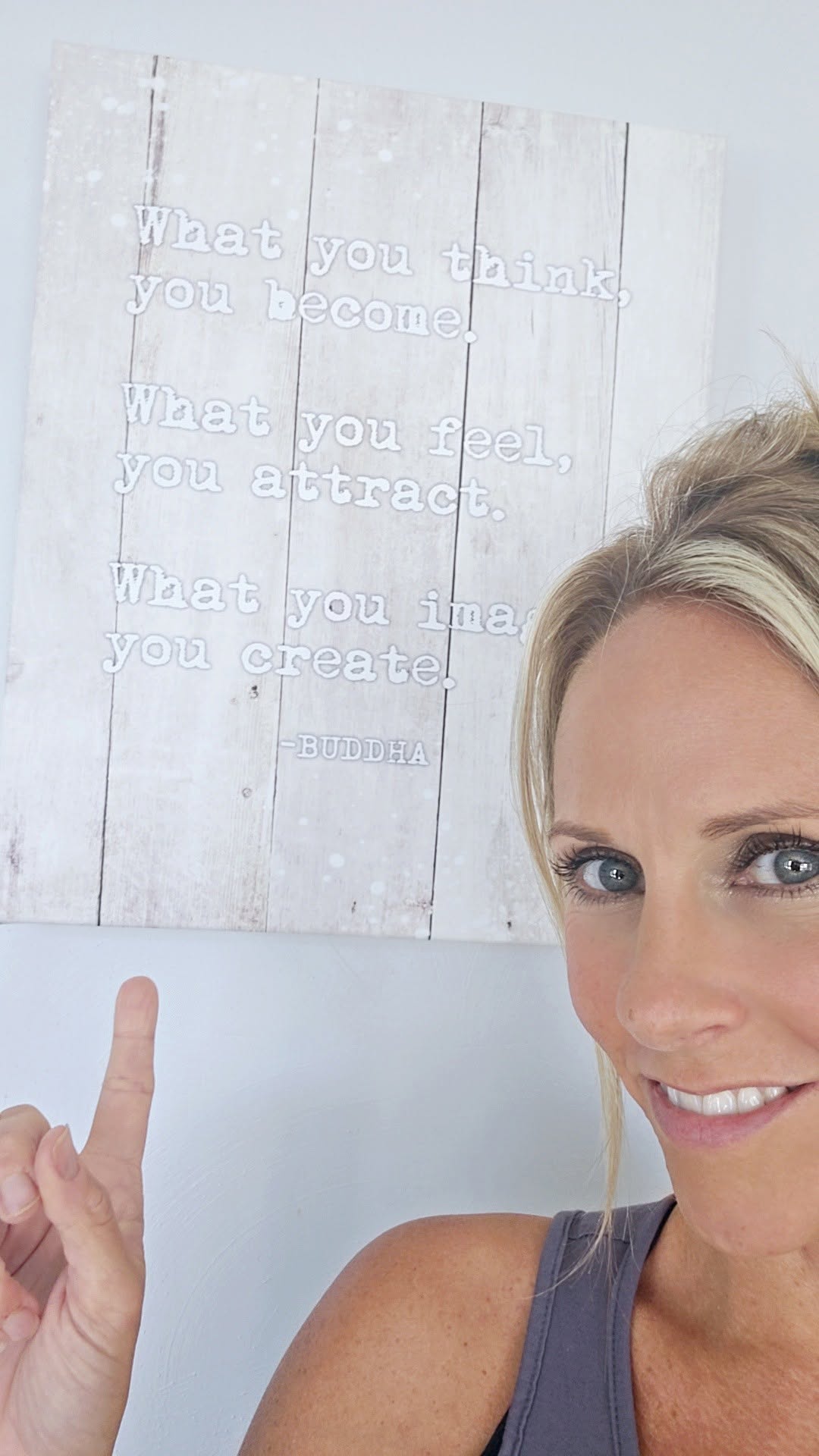 Woman with blonde hair smiling, pointing upward, with a white wall and a quote in the background.