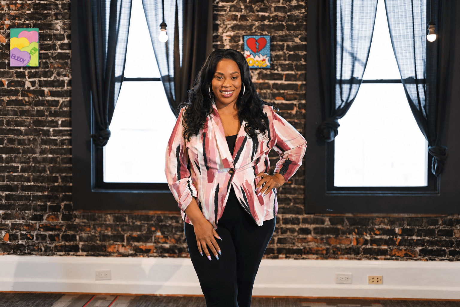 Woman standing in front of brick wall with large windows, smiling, wearing a pink and white patterned blouse and black pants.