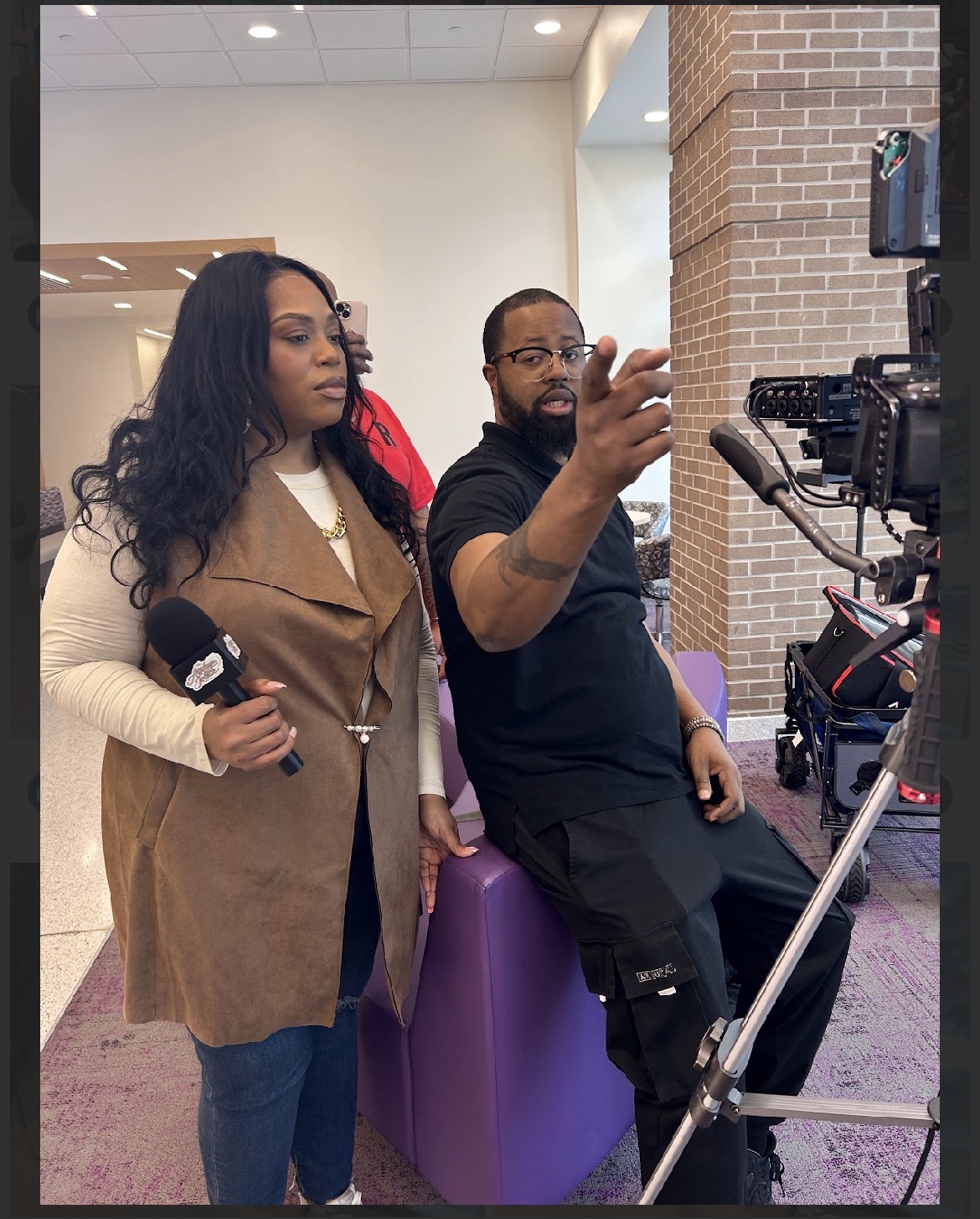 Two people, one seated and one standing, in a room with brick wall, camera equipment, and purple seating.