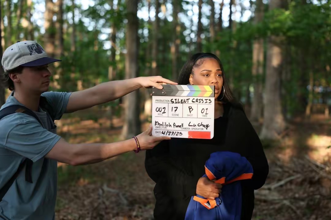 Two people outdoors in a forest, one holding a clapperboard, the other standing nearby, both wearing casual clothing.